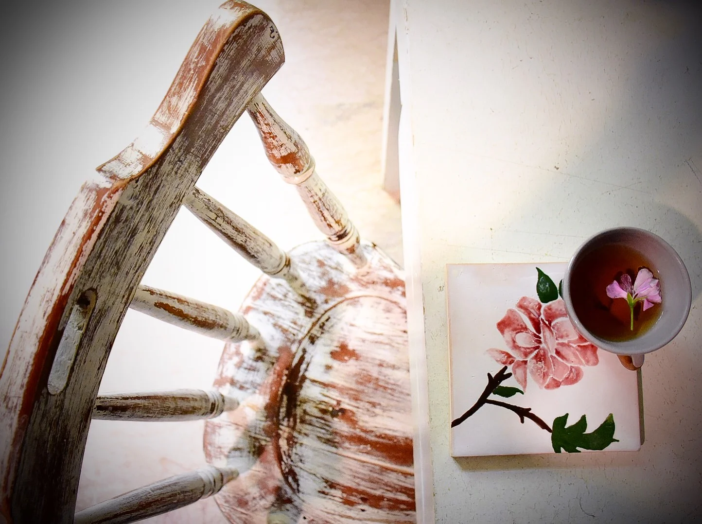 A rustic wooden chair with peeling paint, viewed from above, next to a small table holding a flower-patterned ceramic cup of tea and a pink flower on a watercolor-style floral coaster.