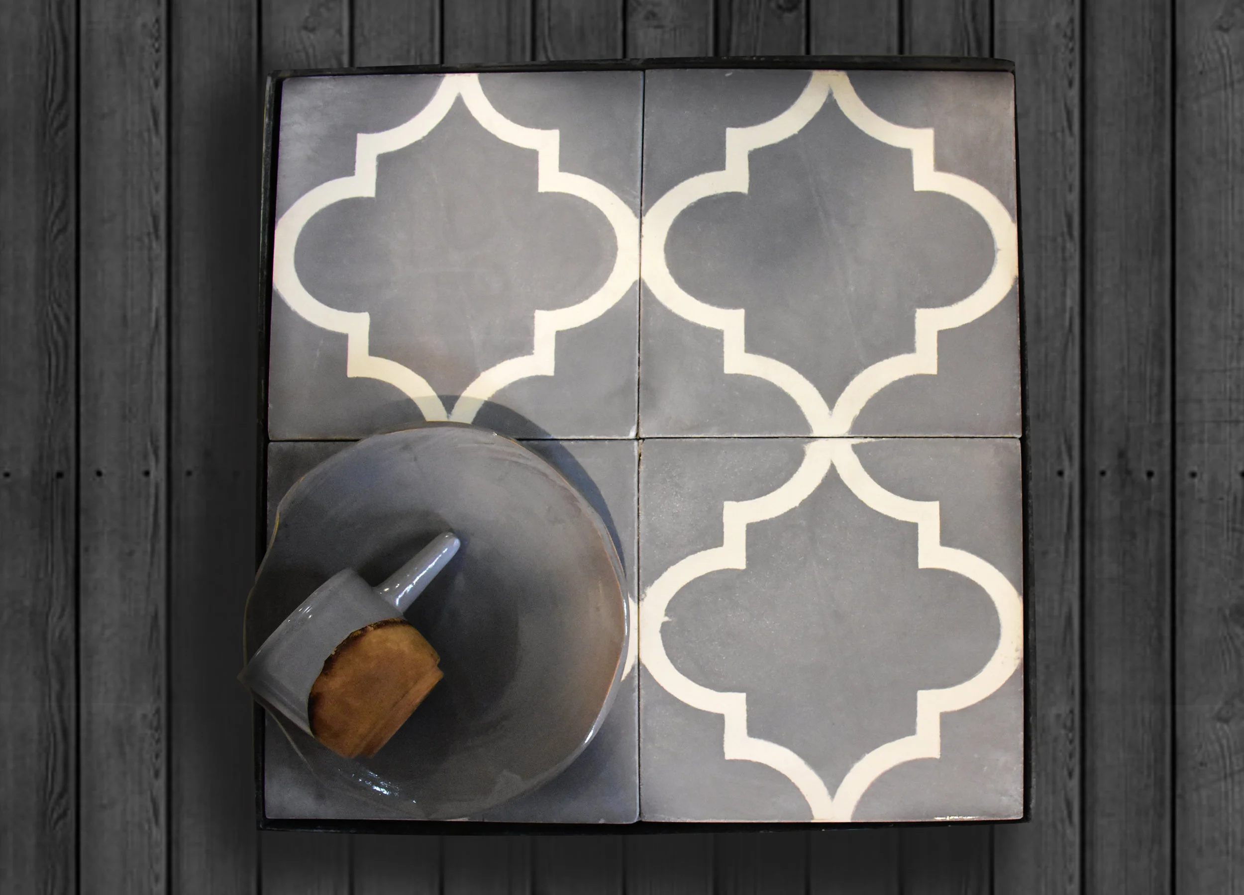 Top-down view of four decorative tiles with a white geometric pattern on a gray background, with a gray mug spilling a small amount of coffee onto the tiles, placed on a dark wooden surface.