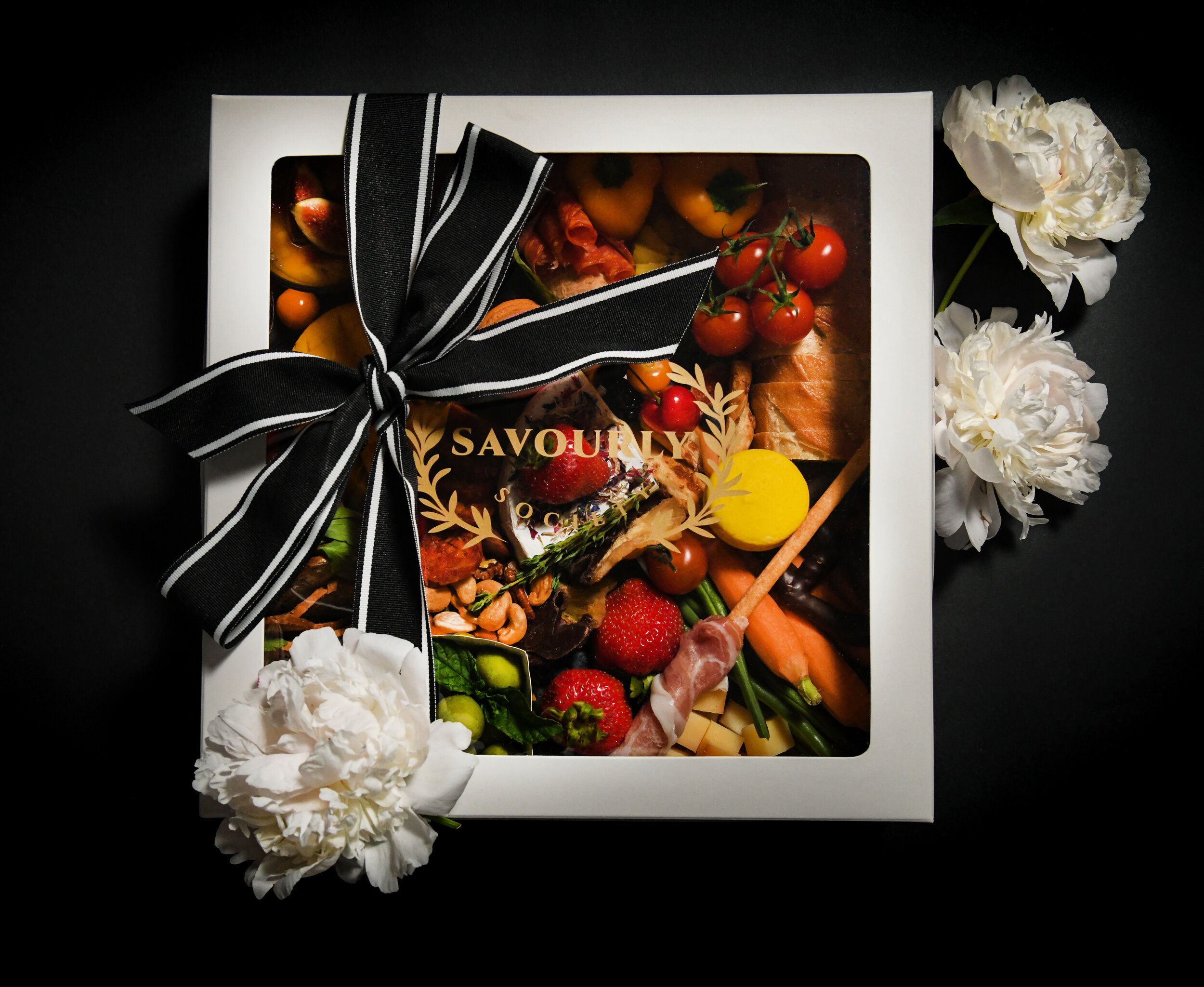 Gift box with assorted foods, tied with a black and white striped ribbon, and decorated with white flowers.