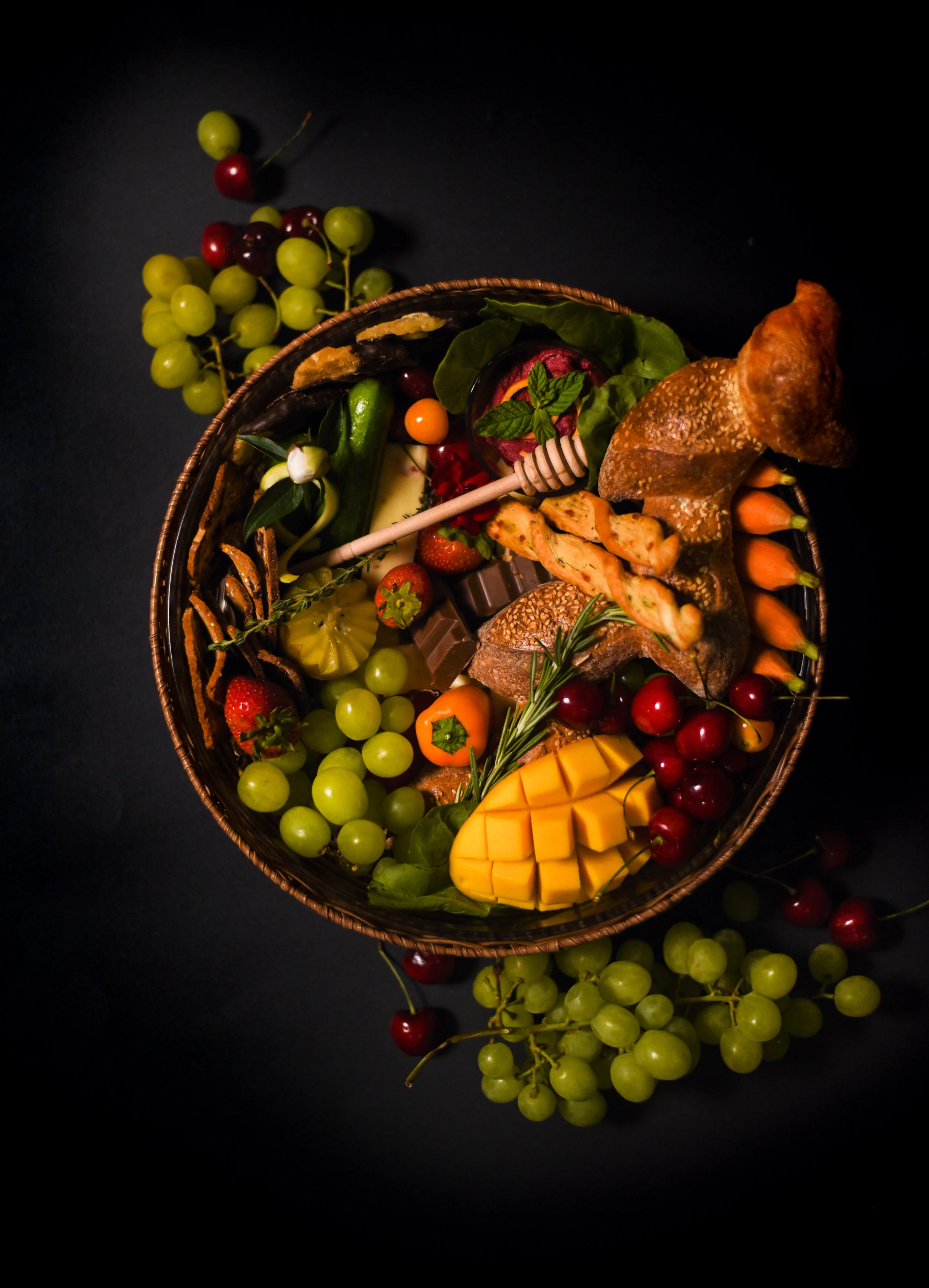 Assorted fruits, cheeses, and snacks in a basket on a black background, including grapes, strawberries, mango, cherry tomatoes, carrots, chocolate, bread, and herbs.