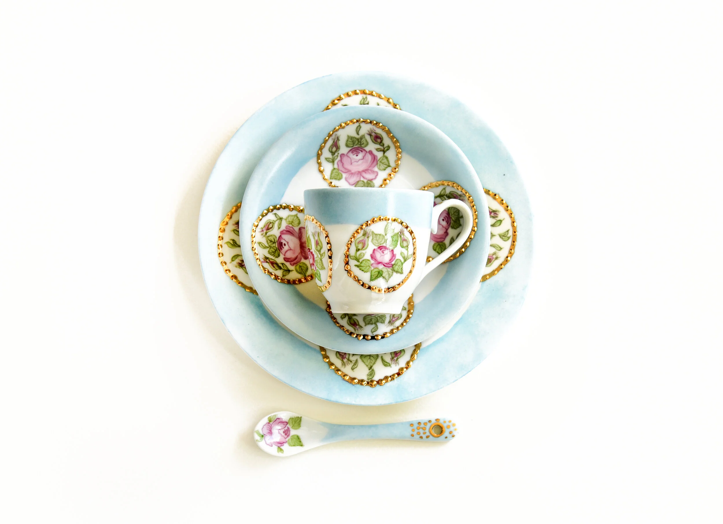 A set of floral-patterned tea cups, saucers, and a spoon with pink roses and green leaves, arranged on a light blue ceramic plate, with a white background.