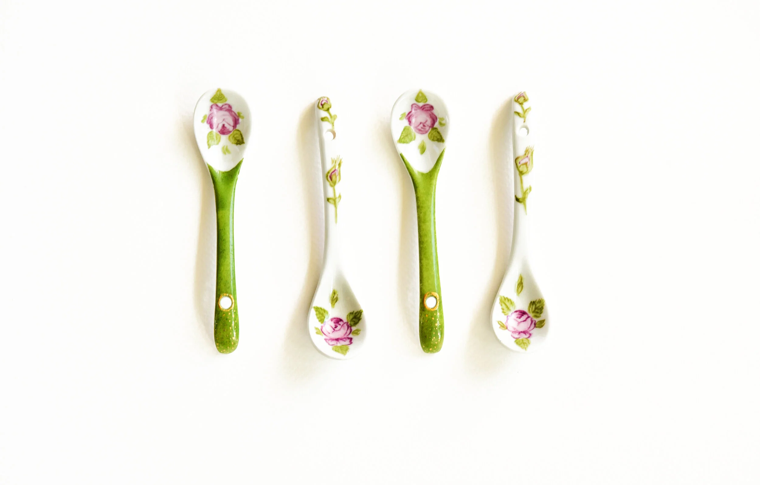 Four porcelain teaspoons with a floral pattern of pink roses and green leaves, two with green handles and two with white handles, arranged on a white surface.