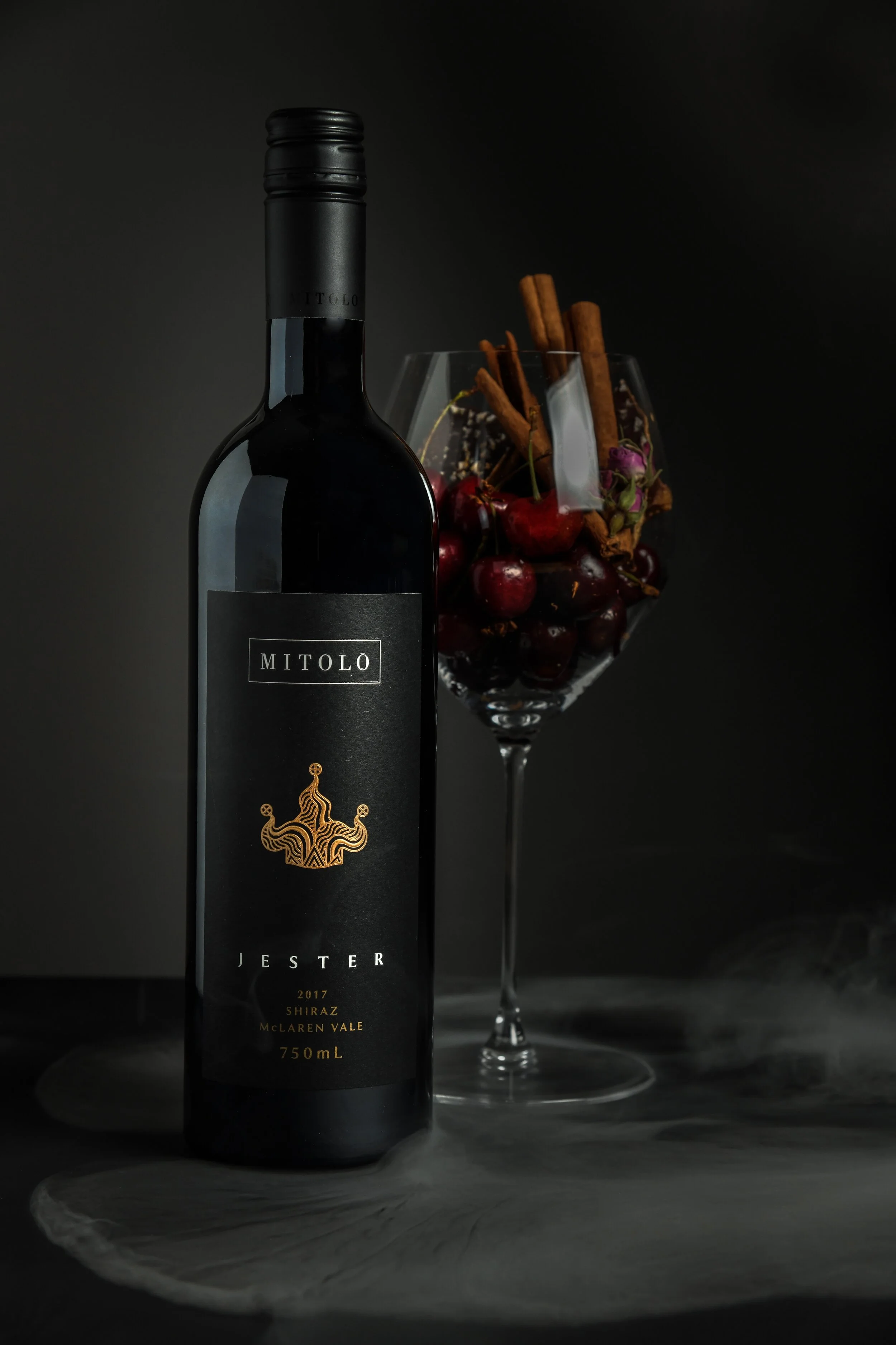A black bottle of Mitolo Jester 2017 Shiraz wine next to a glass filled with cherries and cinnamon sticks, on a dark surface with a dark background.
