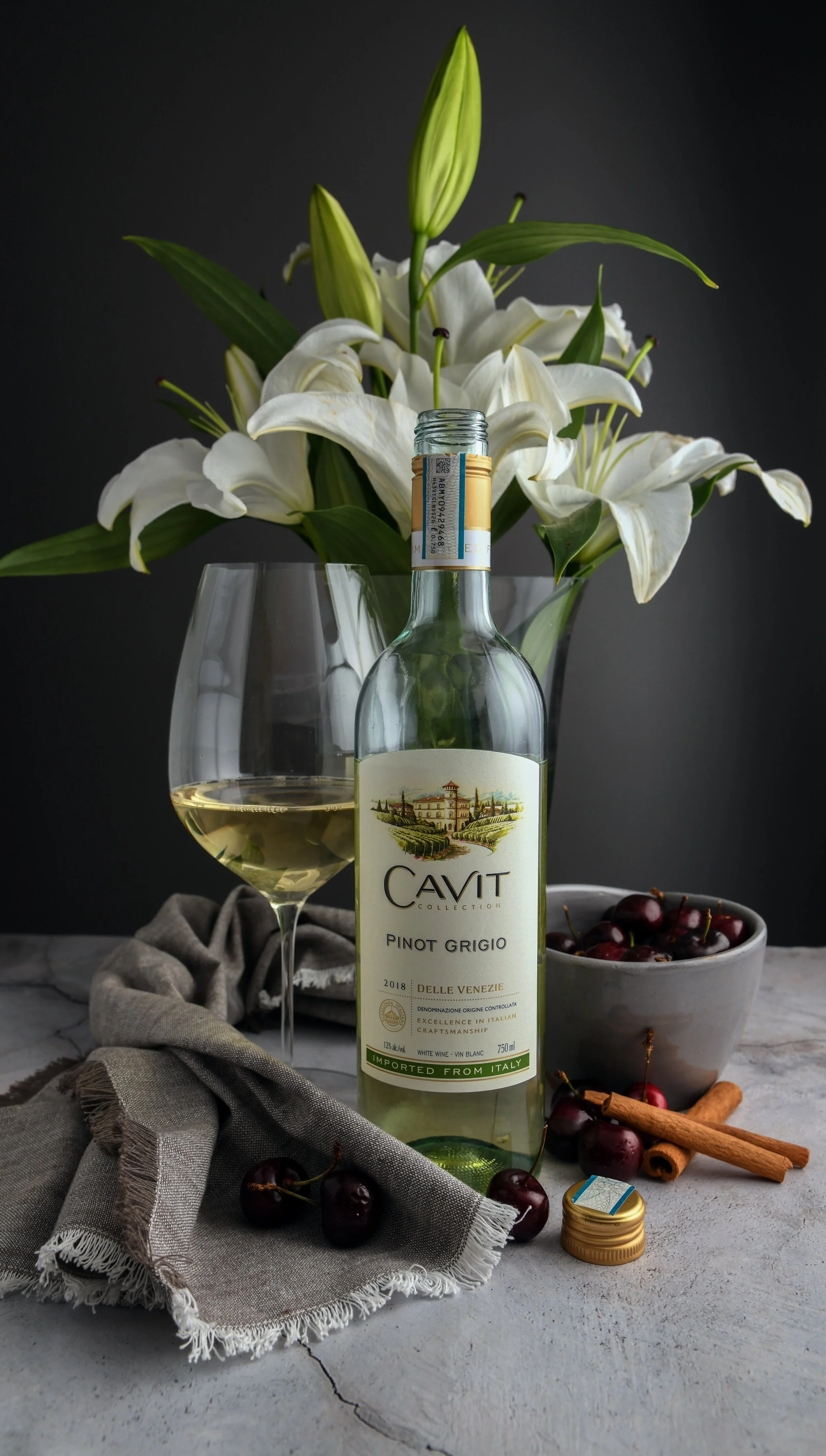 A bottle of Cavit Pinot Grigio wine with a glass of white wine beside it, a bowl of cherries, cinnamon sticks, and white lilies in a vase on a gray surface against a dark background, with a gray cloth under the cherries and wine glass.