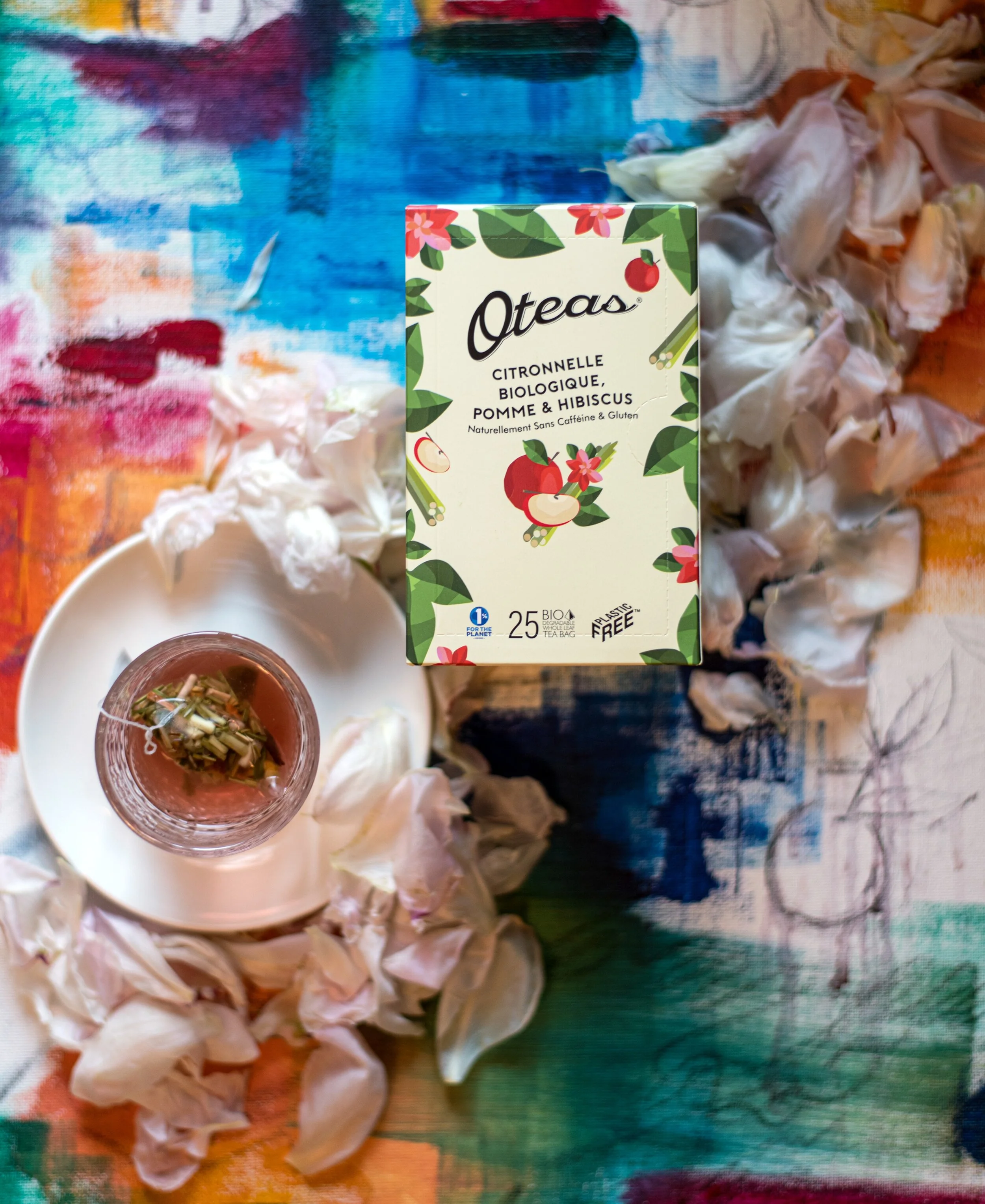 A box of Otea citronnelle biologique tea with apple and hibiscus flavors, placed on a colorful, abstract painted table. Surrounding the box are crumpled tea bags and a glass teacup with a tea infuser, on a white saucer.