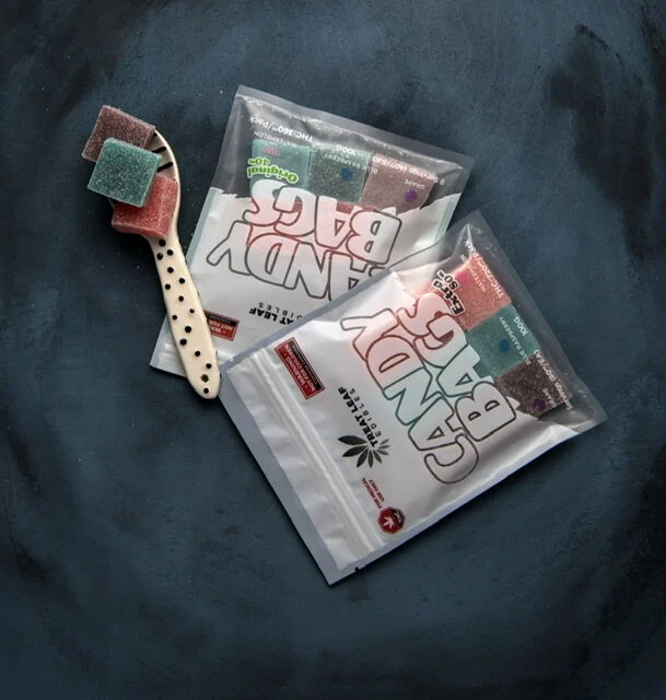 Two unopened packages of CBD Gummies with a white spatula containing colorful gummy candies, set on a dark surface.