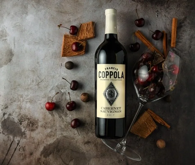 A bottle of Coppola Cabernet Sauvignon wine, a wine glass filled with cherries, and scattered cherries, cinnamon sticks, and corks on a textured gray surface.