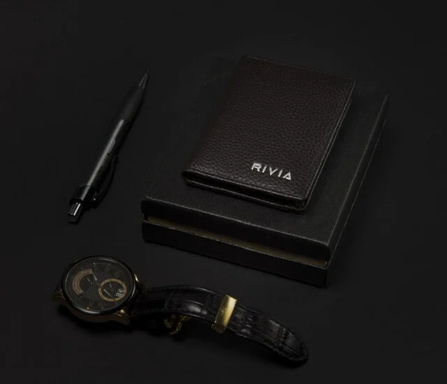 Black pen, leather wallet with 'RIVIA' branding, two black notebooks, and a wristwatch with a black leather strap on a black background.