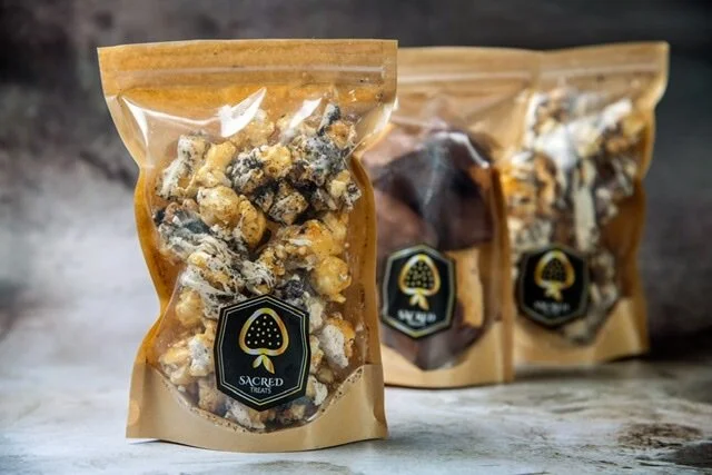 Three resealable bags of assorted snack items with a black and gold Sacred Herb label, placed on a grey surface with a dark background.