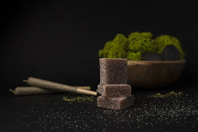 Stack of brown sugar cubes, green wasabi in a wooden bowl, two beige sticks, black background, scattered sugar crystals.