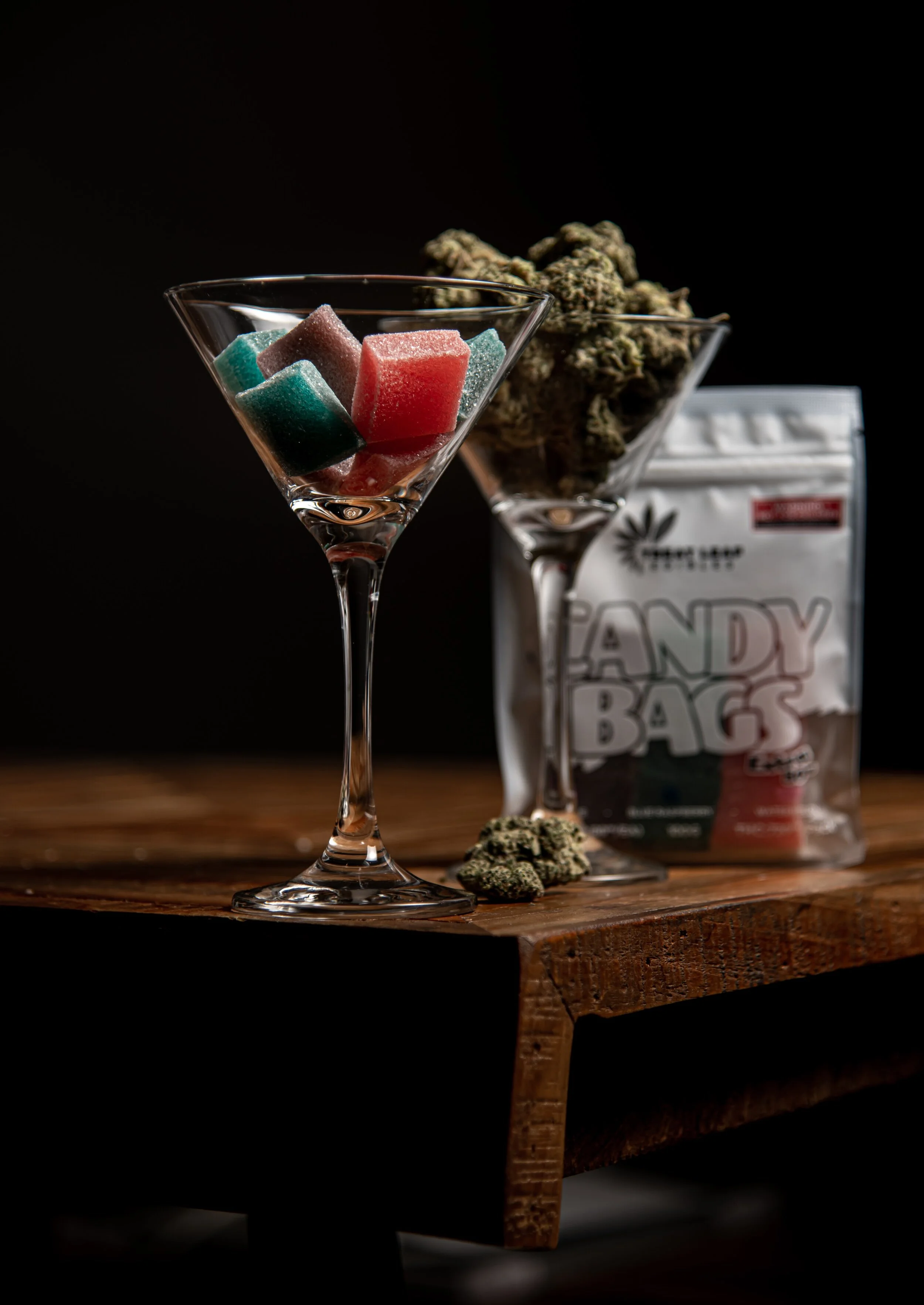 Two martini glasses, one filled with colorful gummy candies, the other with cannabis buds, placed on a wooden surface with a package of candy in the background and cannabis buds scattered around.