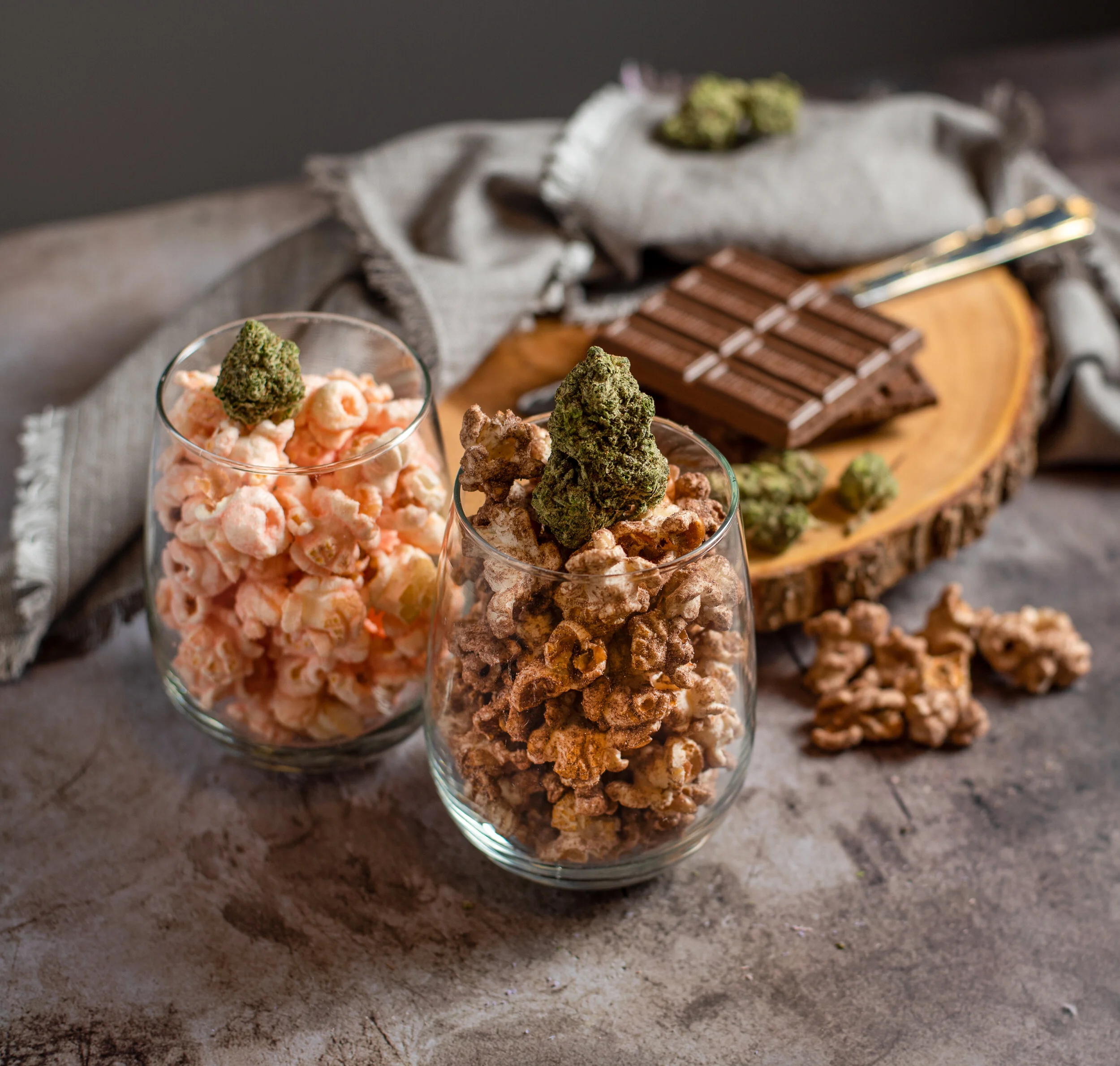 Two glasses filled with popcorn topped with cannabis buds, with chocolate bars and additional cannabis buds on a wooden slice in the background.