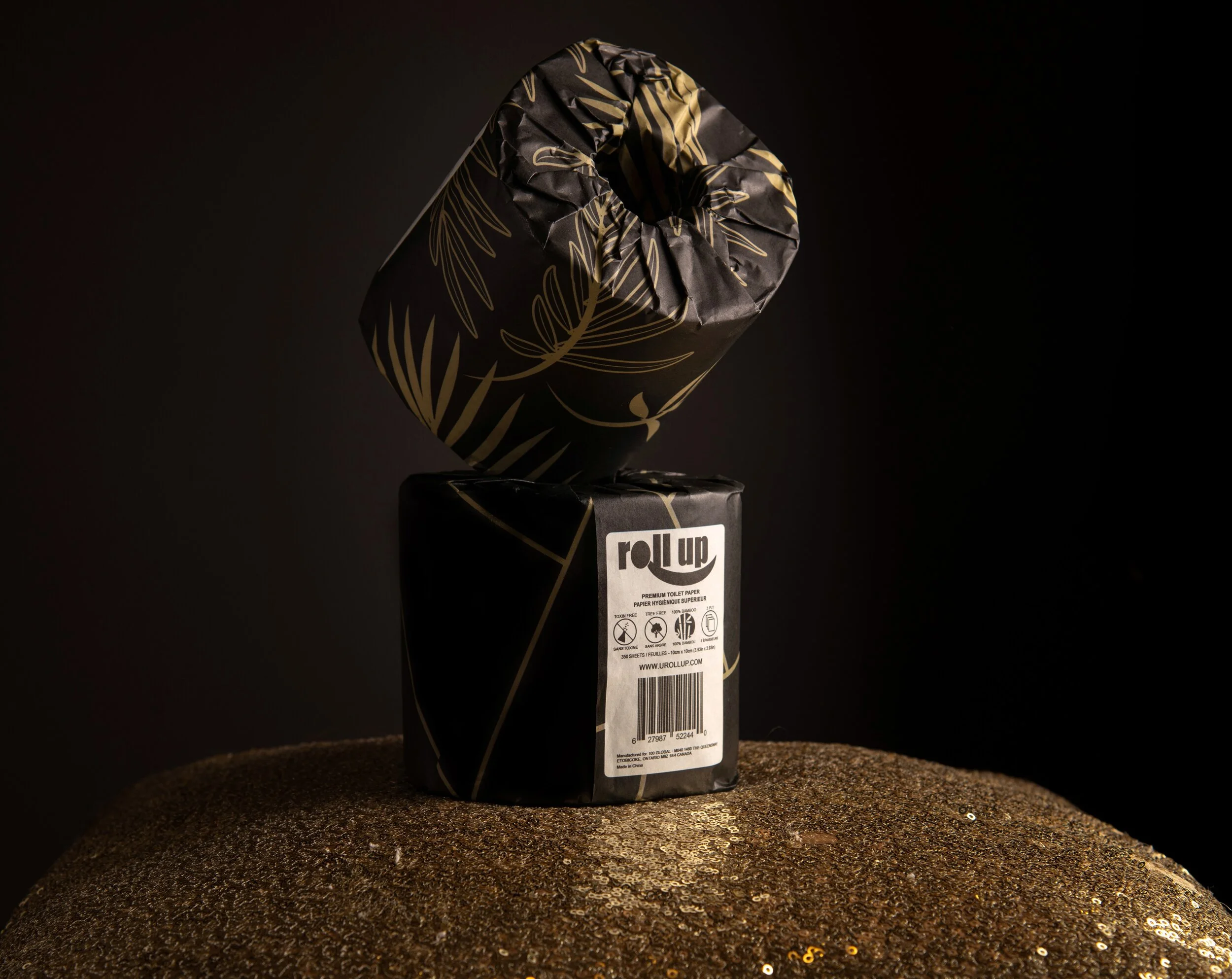 Stack of two black toilet paper packages, with a top package partially unwrapped and resting diagonally on the lower package, on a brown sparkling surface against a dark background.