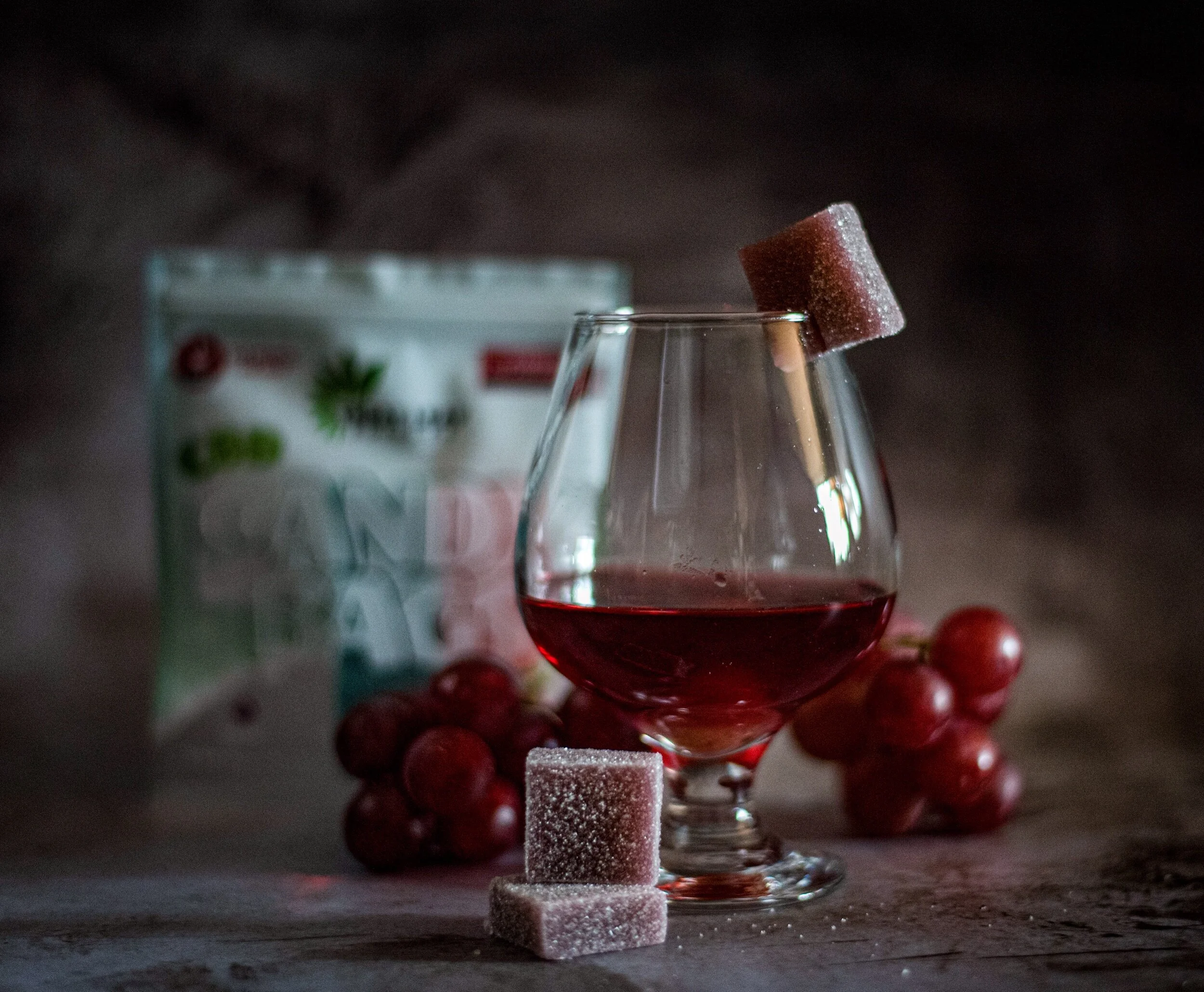 A glass of red wine with a sugar cube on the rim, surrounded by grapes and sugar cubes, with a package in the background.