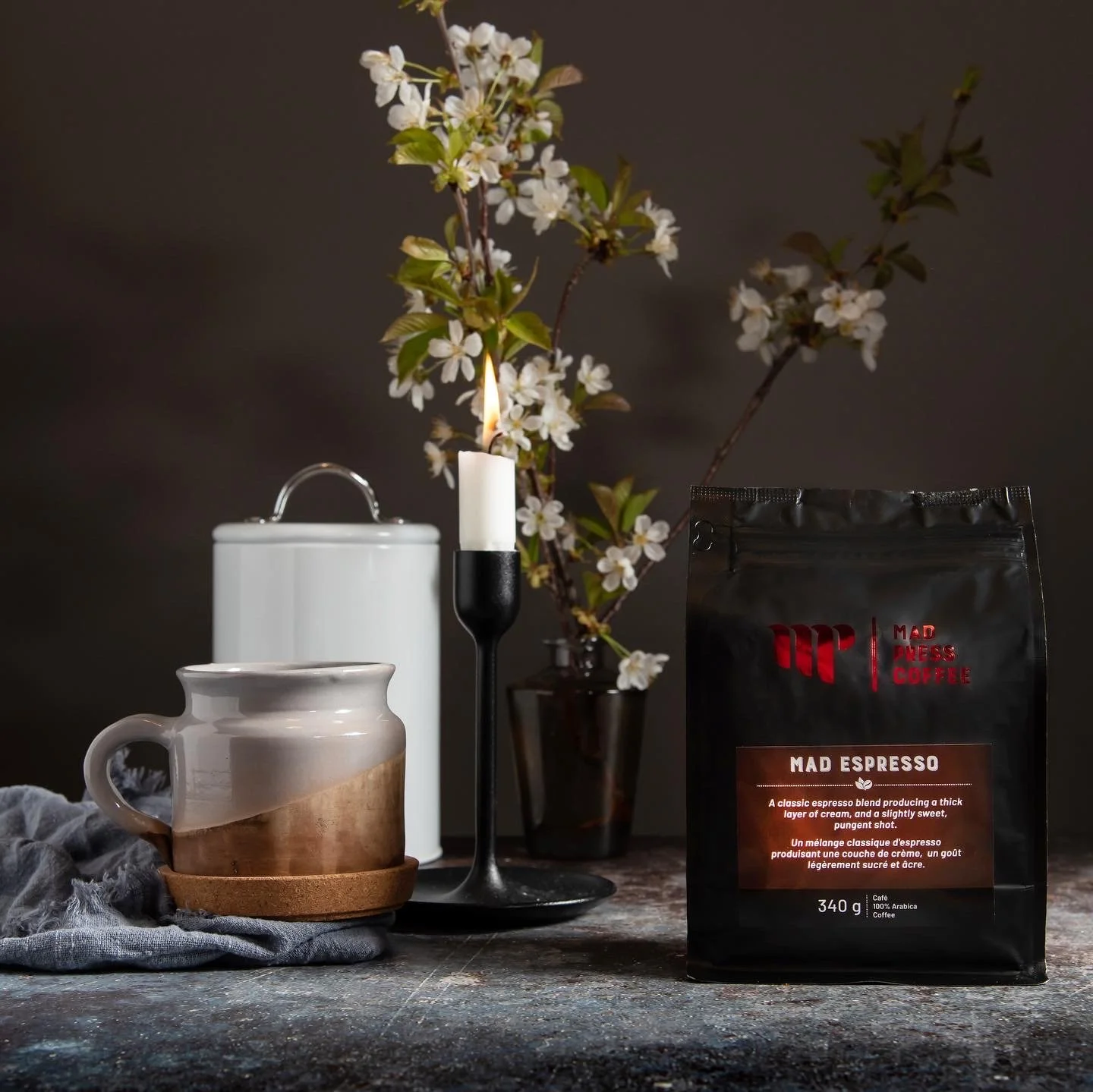 A bag of Mad Espresso coffee beans, a ceramic mug on a wooden coaster, a white container, a black candle holder with a candle, and a vase with white blossoms on a dark textured surface.