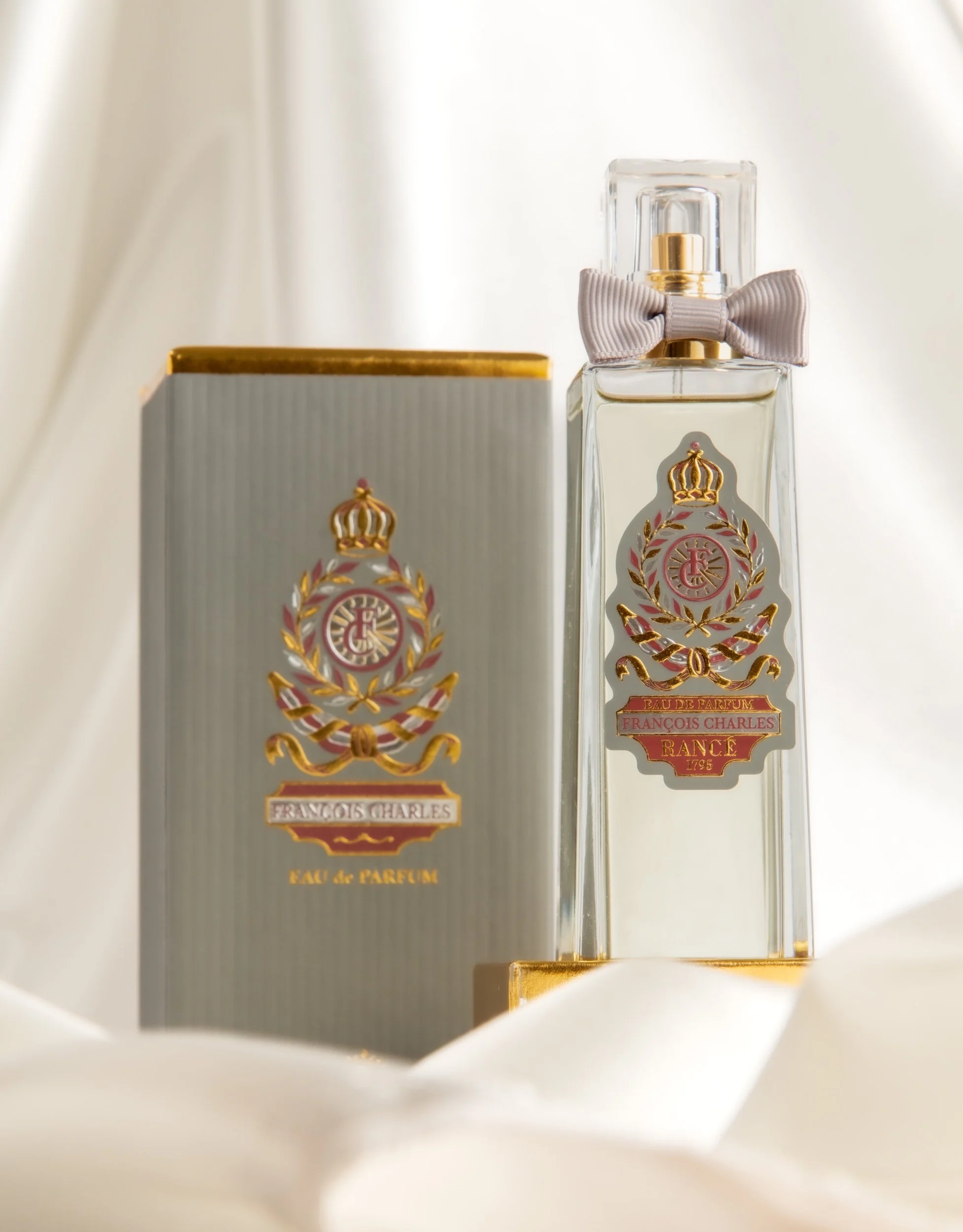 A perfume bottle with a gold crown logo and pink label, accompanied by its matching box, displayed on white fabric with soft lighting.