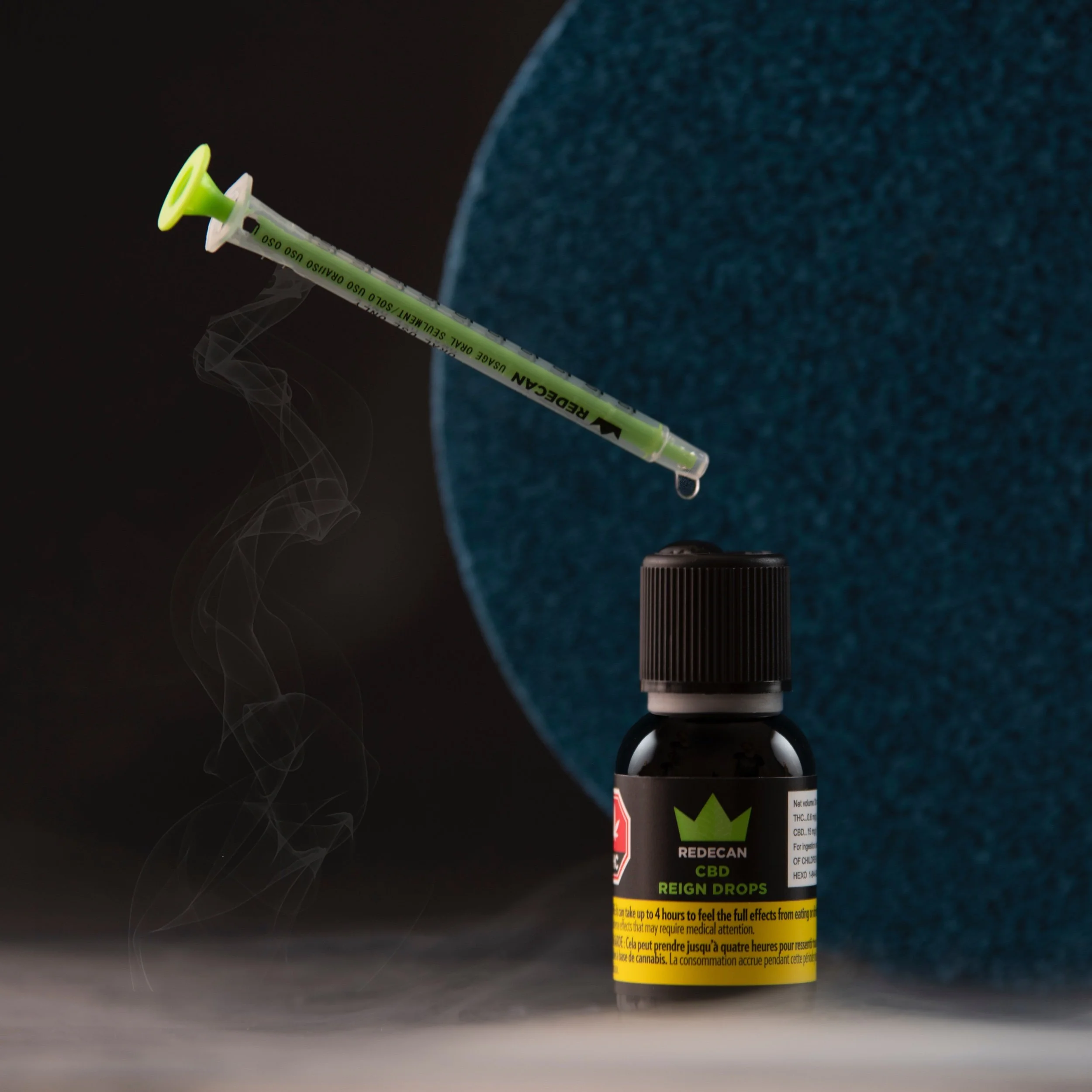 A vape cartridge with a black cap and a label reading 'REDECAN CBD REIGN DROPS' is placed on a surface. A syringe with a green needle tip and a yellow warning label is positioned above, with a drop of liquid falling from it. Smoke wisps are visible.