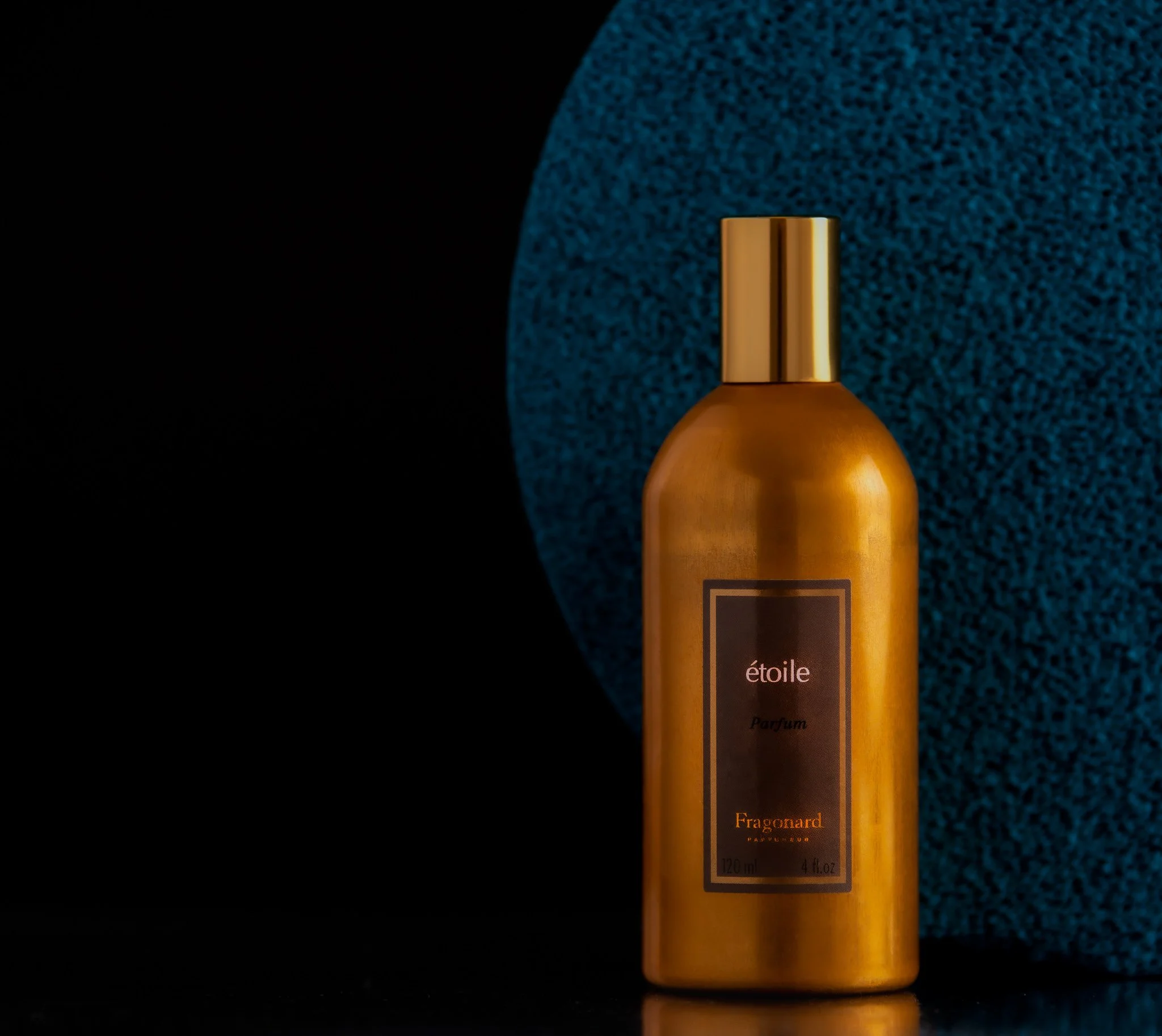 A gold-colored bottle of perfume labeled 'étoile Parfum' stands in front of a textured blue circle with a black background.
