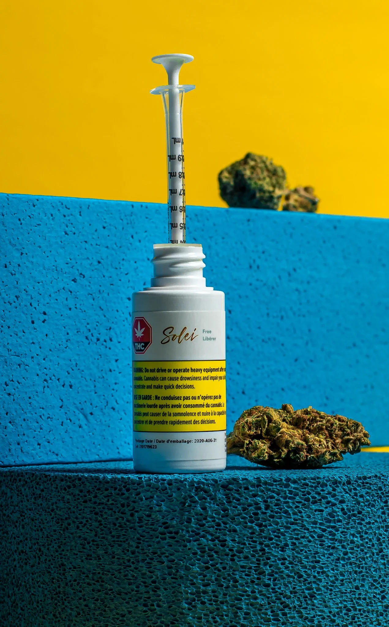 A white medical bottle labeled 'Solei' with a THC warning symbol, a yellow caution strip, and a dosage syringe inserted into the bottle. A cannabis bud is placed beside the bottle on a textured blue surface with a blue and yellow background and a can