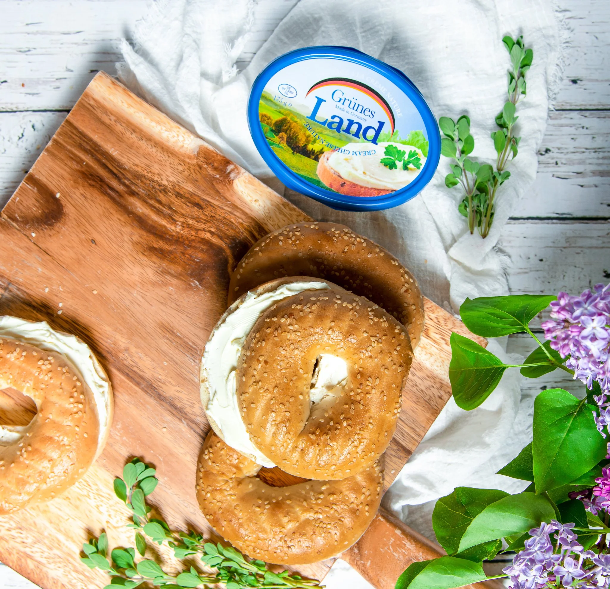 Bagels with cream cheese on a wooden cutting board, a container of cream cheese labeled "Grünes Land," and purple lilac flowers on a white surface.