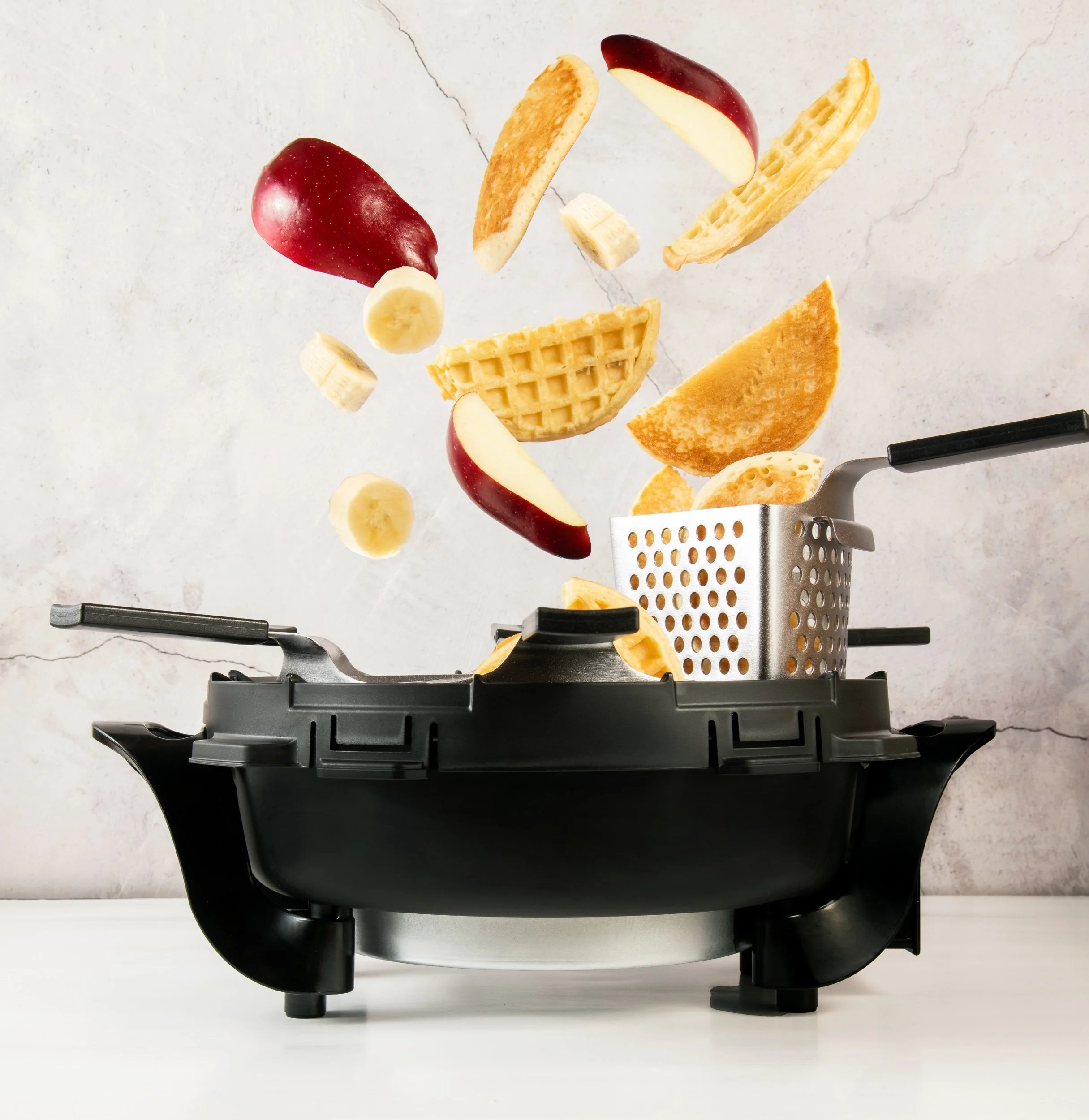 Slices of bananas, apple, waffle, pancake, and waffle cone falling into a black air fryer on a white surface against a light textured background.