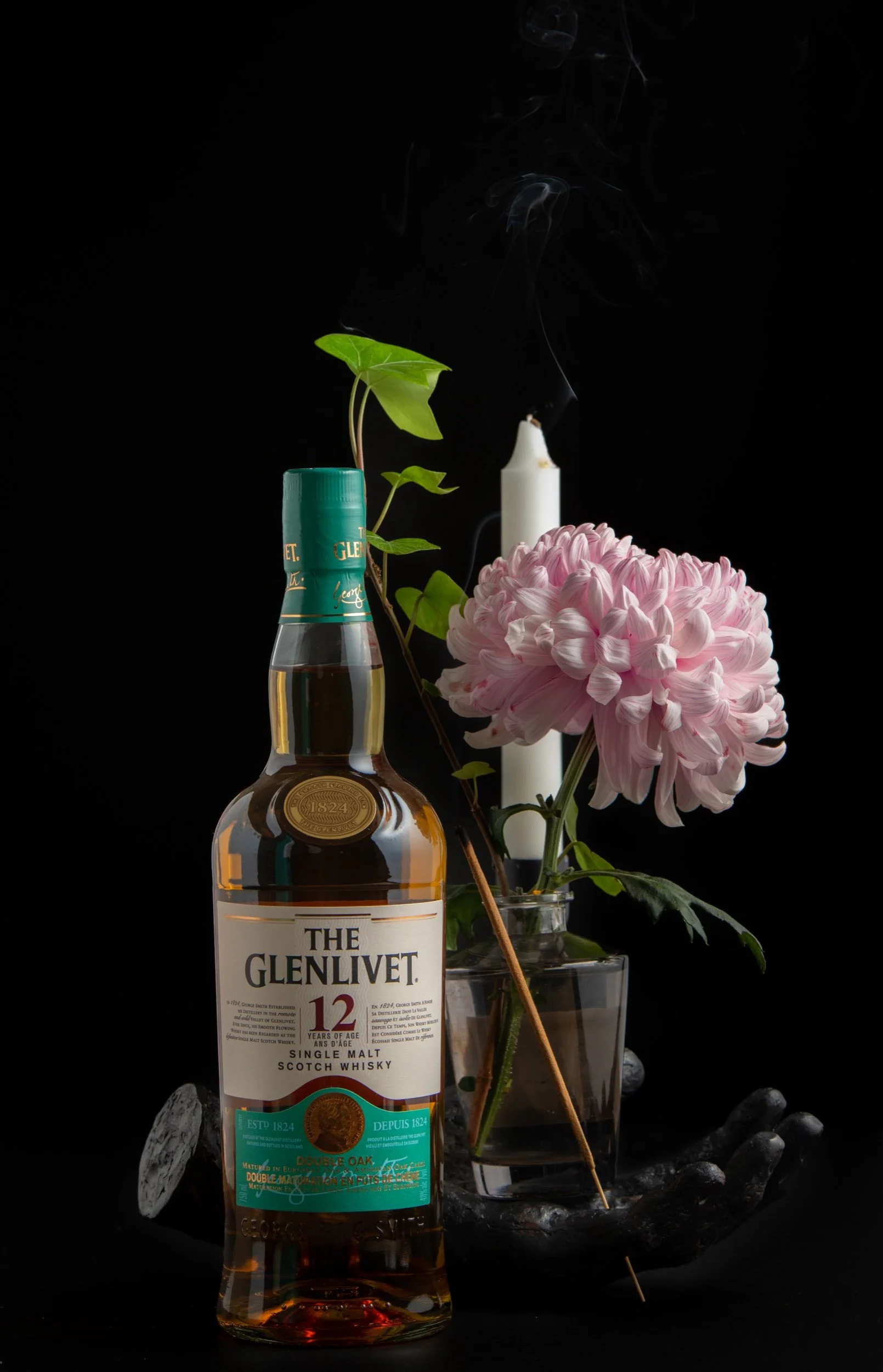 A bottle of Glenlivet 12-year-old single malt scotch whisky, a pink chrysanthemum flower in a glass vase, a white candle, and a smoke trail against a black background.