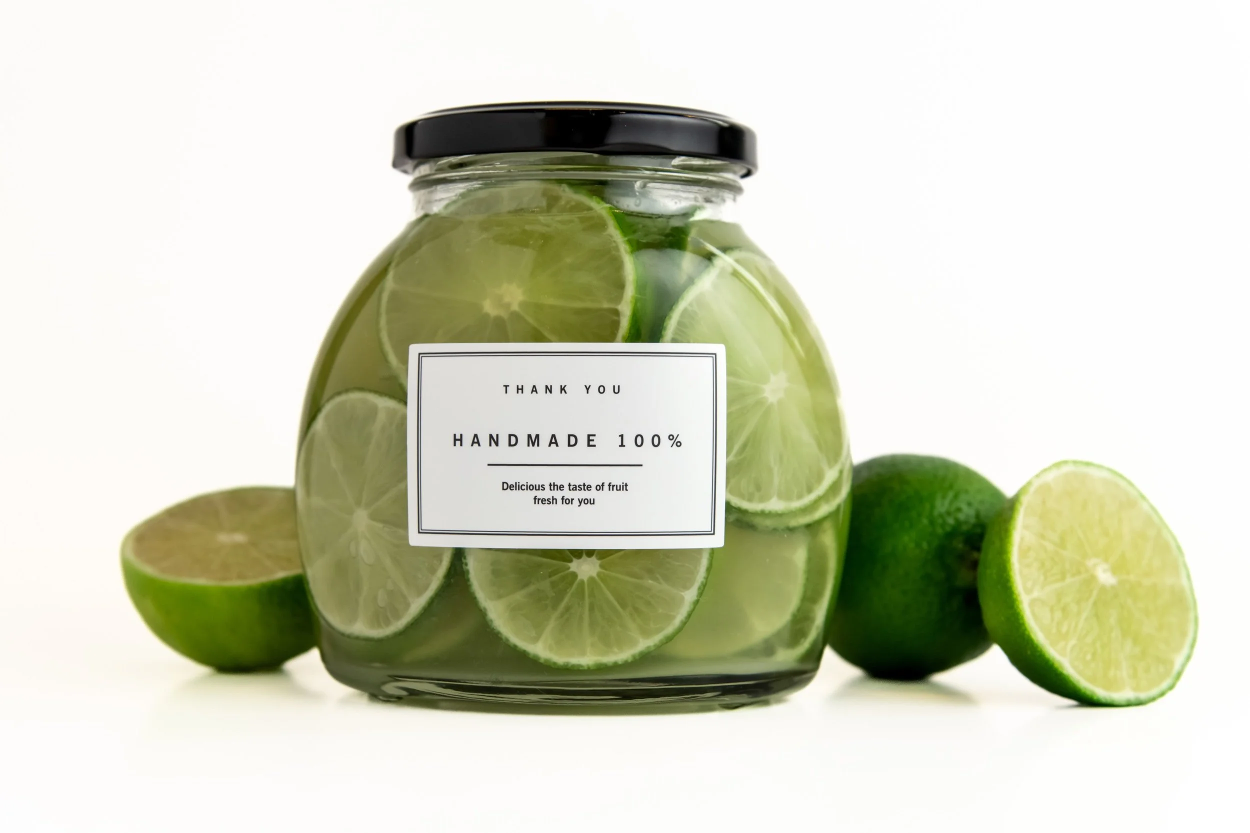 A glass jar filled with sliced limes and lemon slices, with a label that says "Thank You Handmade 100%, Delicious the taste of fruit fresh for you," surrounded by additional whole and halved limes on a white background.