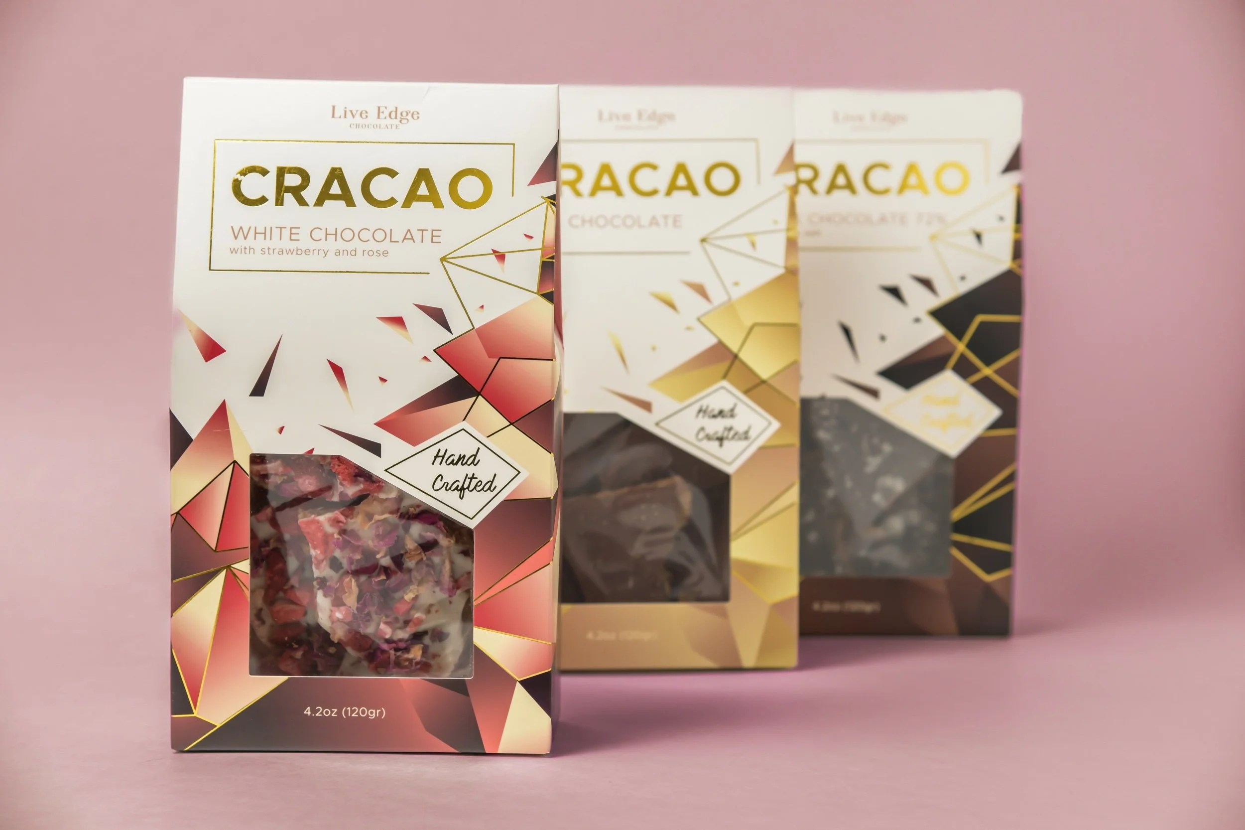 Three boxes of CRACAO chocolate with geometric design, labeled as white, dark, and mixed chocolates, on a pink background.