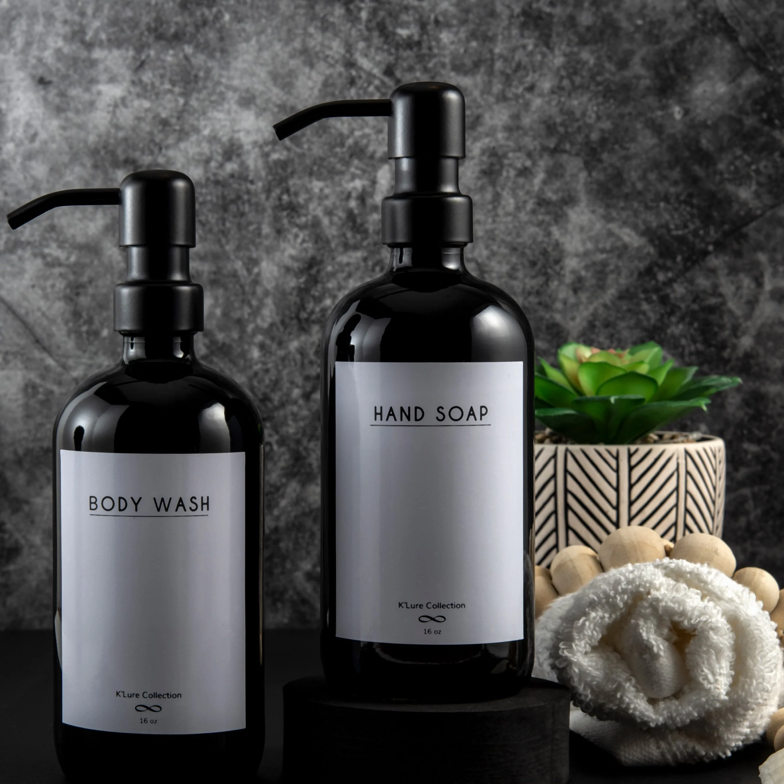 Black bottles of body wash and hand soap placed on a black surface, with a potted succulent plant and a rolled white towel nearby.