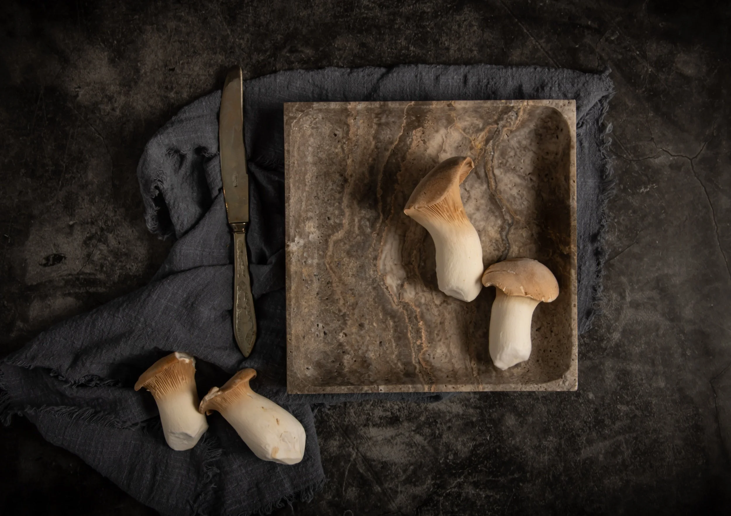 Four white mushrooms with brown caps on a marble cutting board, with a gray cloth, a vintage butter knife, and a dark textured surface surrounding it.