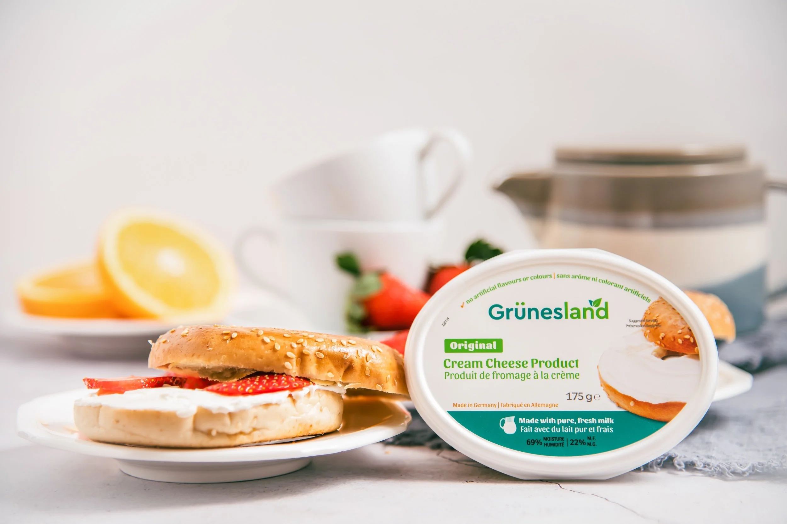 Cream cheese spread on a bagel with strawberries, with a container of Grünenland cream cheese in the foreground and strawberries, lemon slices, tea cups, and a sugar container in the background.