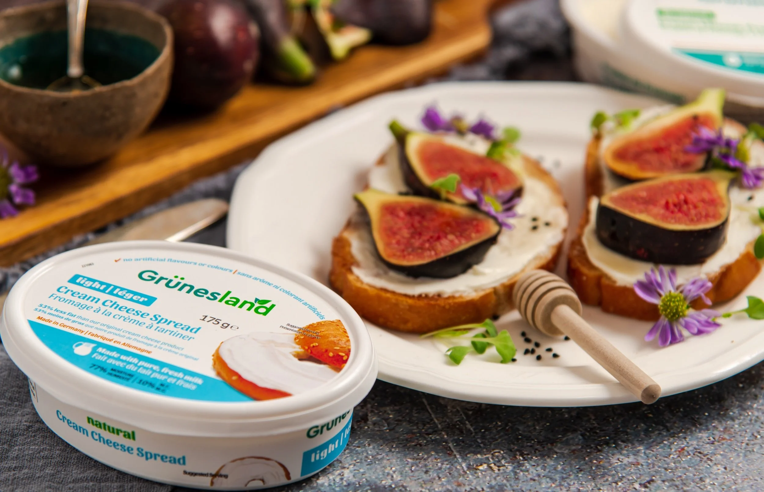 Open container of Grüneland light layer cream cheese spread, with slices of fig and dollops of cream cheese on toasted bread topped with purple edible flowers, on a white plate. In the background, a wooden board with more figs and a bowl of jam.