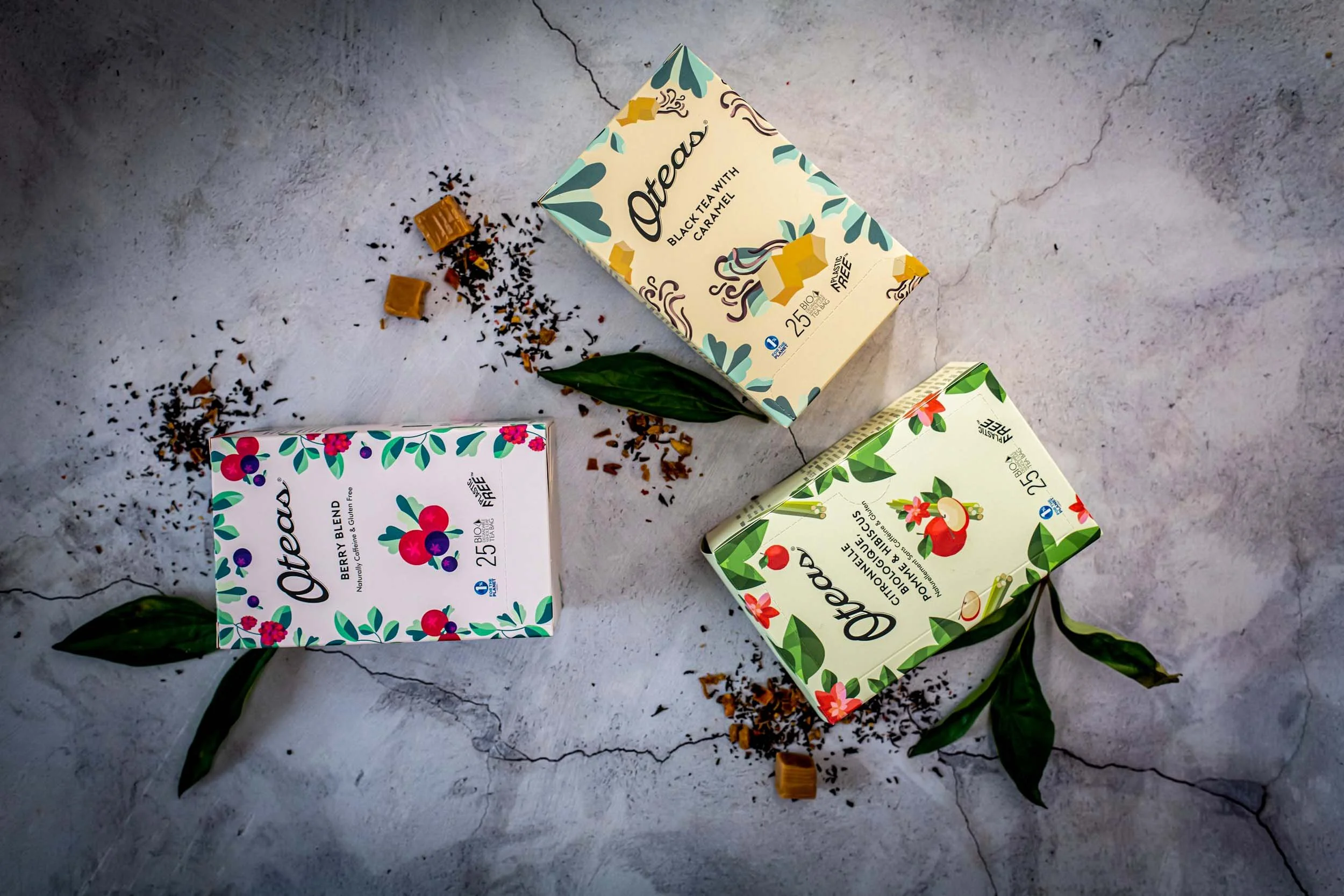 Three boxes of Oateas tea with loose tea leaves and ingredients scattered around, garnished with green leaves on a concrete surface.