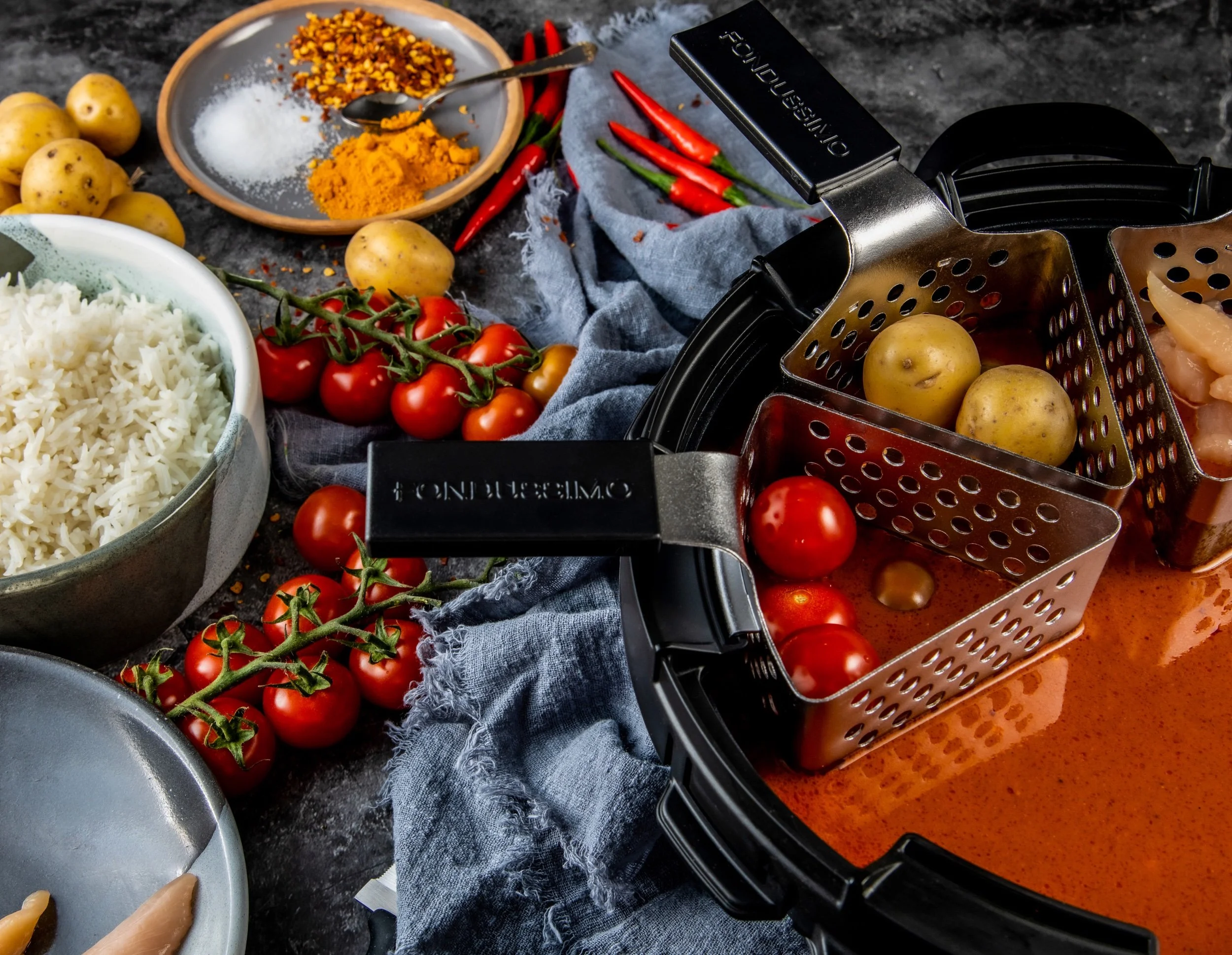 A kitchen countertop with ingredients including cherry tomatoes on the vine, potatoes, rice, spices, chili peppers, and a pot of tomato sauce, with an air fryer in the background.