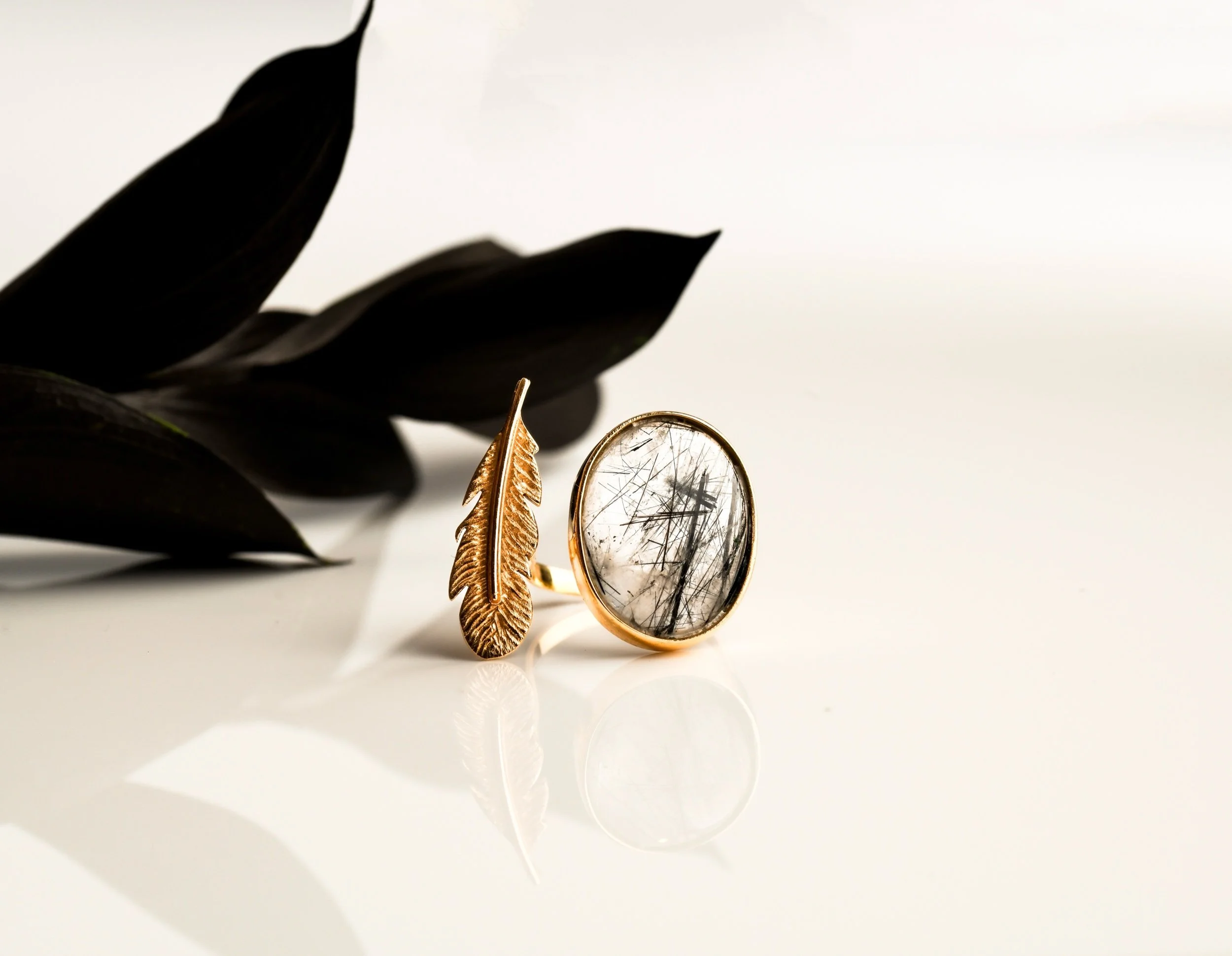 Gold ring with oval clear gemstone containing black and gray hair-like inclusions, and a gold feather-shaped ring, placed on a white surface with black leaves in the background.