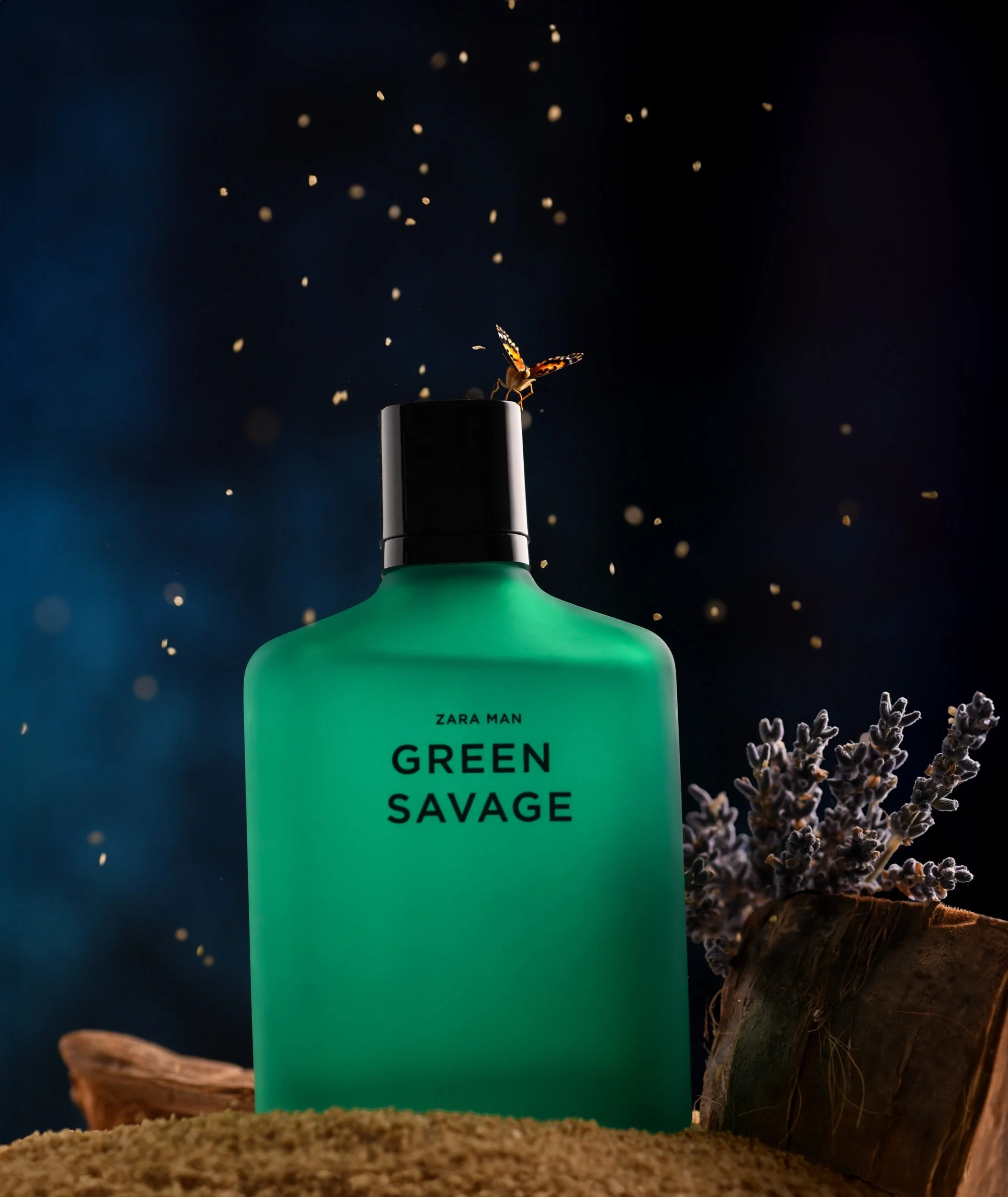 A green bottle of Zara Man Green Savage cologne placed on beige rough surface with lavender flowers and wood pieces beside it, and a butterfly perched on the bottle's cap, with floating particles in the dark background.