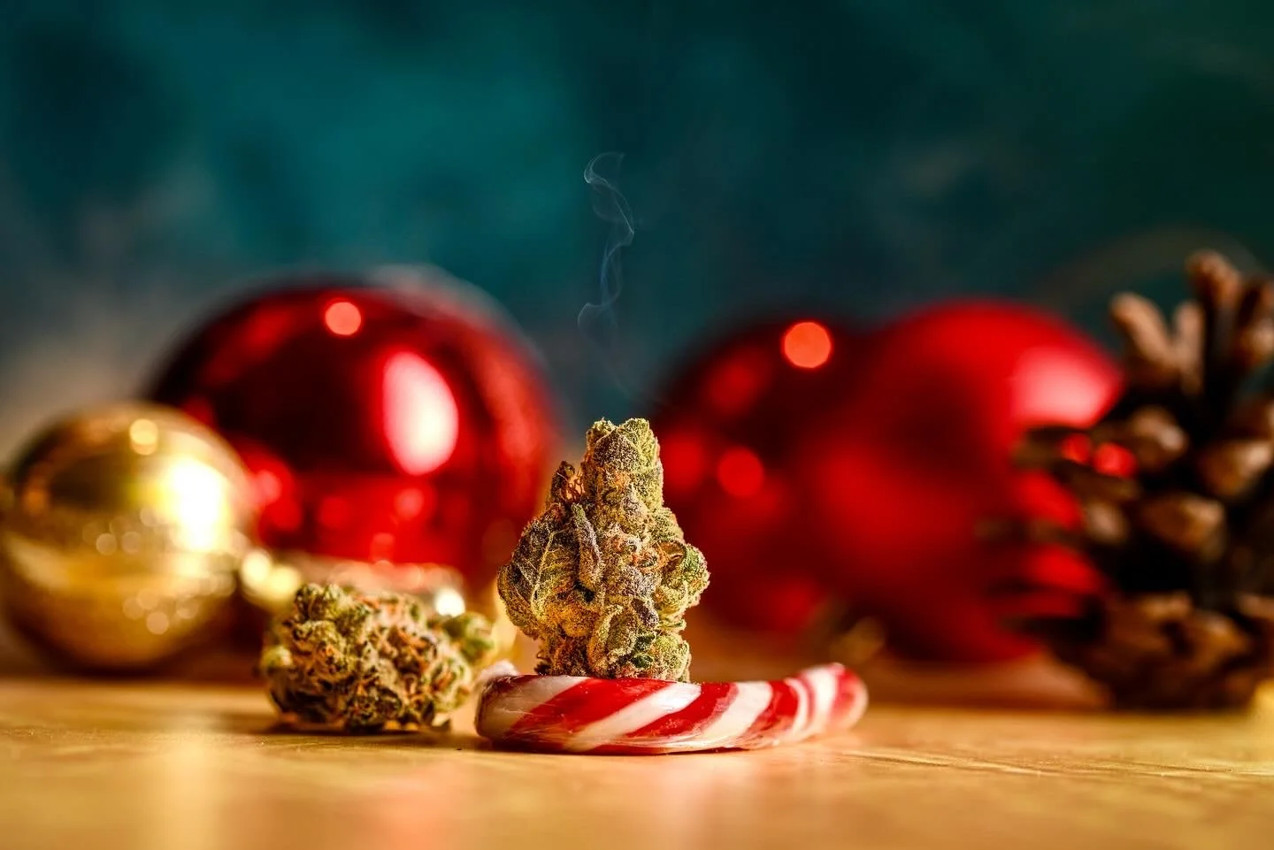 Cannabis buds, a peppermint candy cane, Christmas ornaments, and pine cone on a wooden surface.