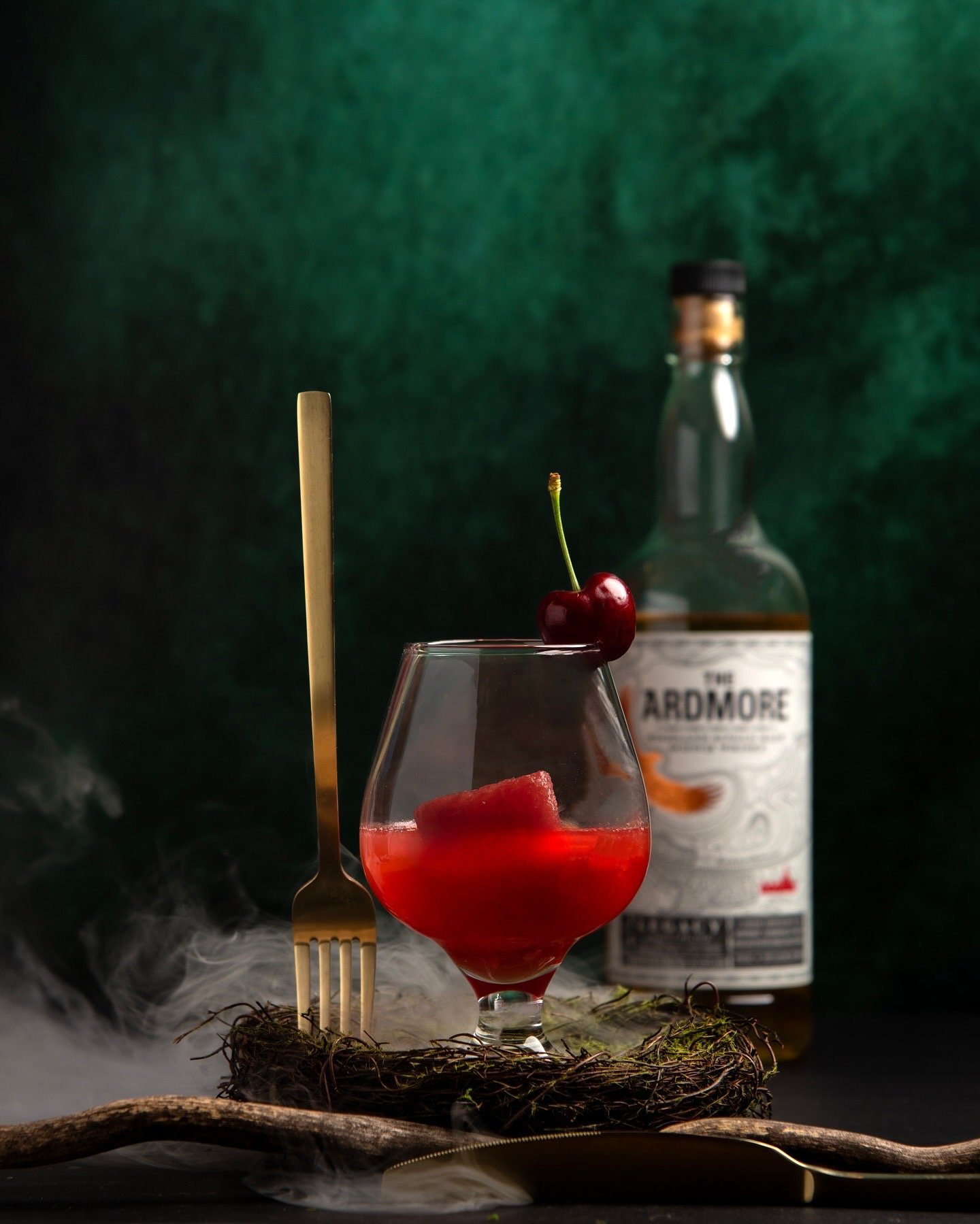 A glass with a red cocktail garnished with a cherry and a cherry slice, a gold fork, a bottle of Ardmore whiskey in the background, and some mist and twigs at the base.