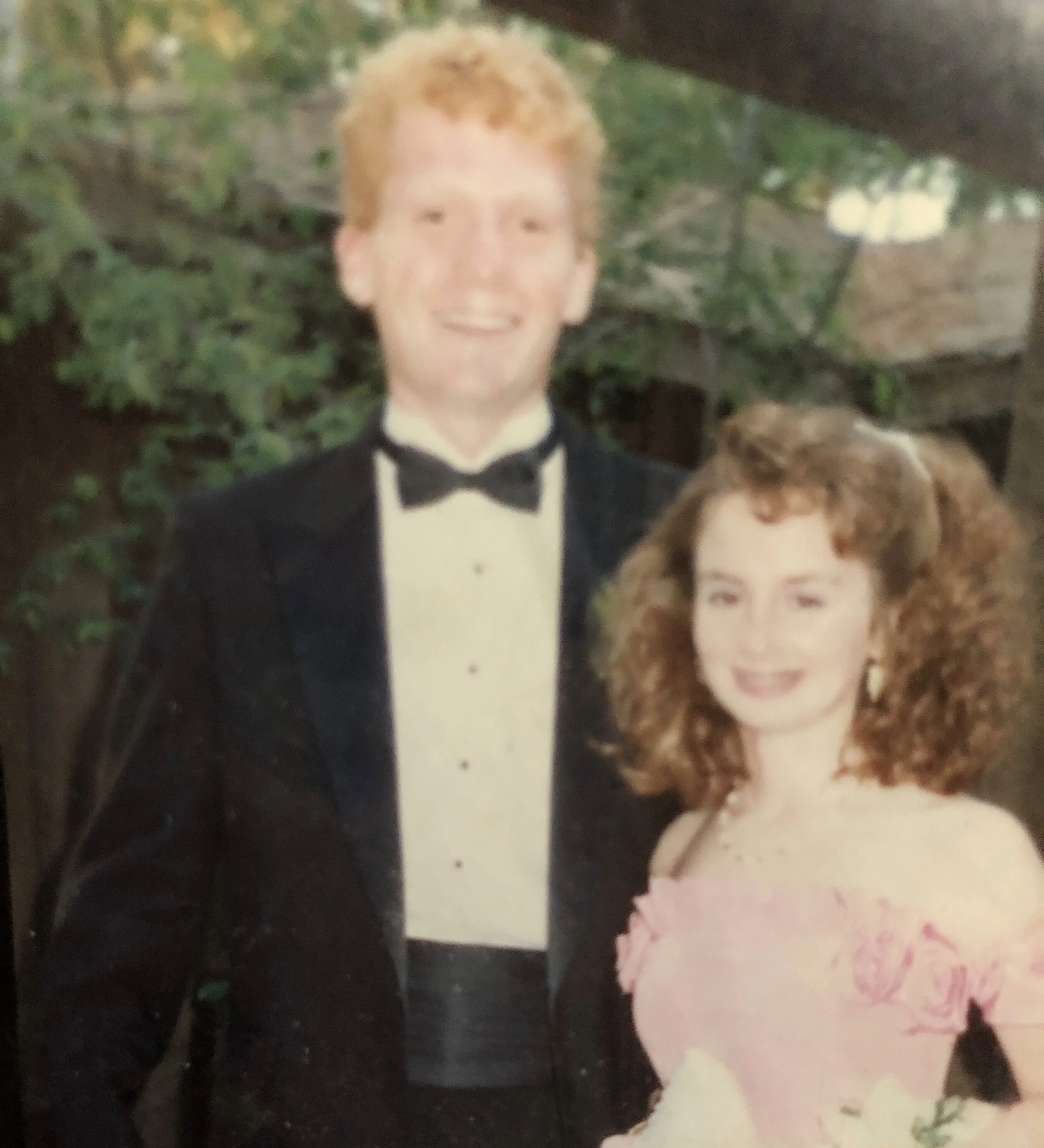 Young couple in formal wear, man in tuxedo, woman in floral pink dress, smiling outdoors.