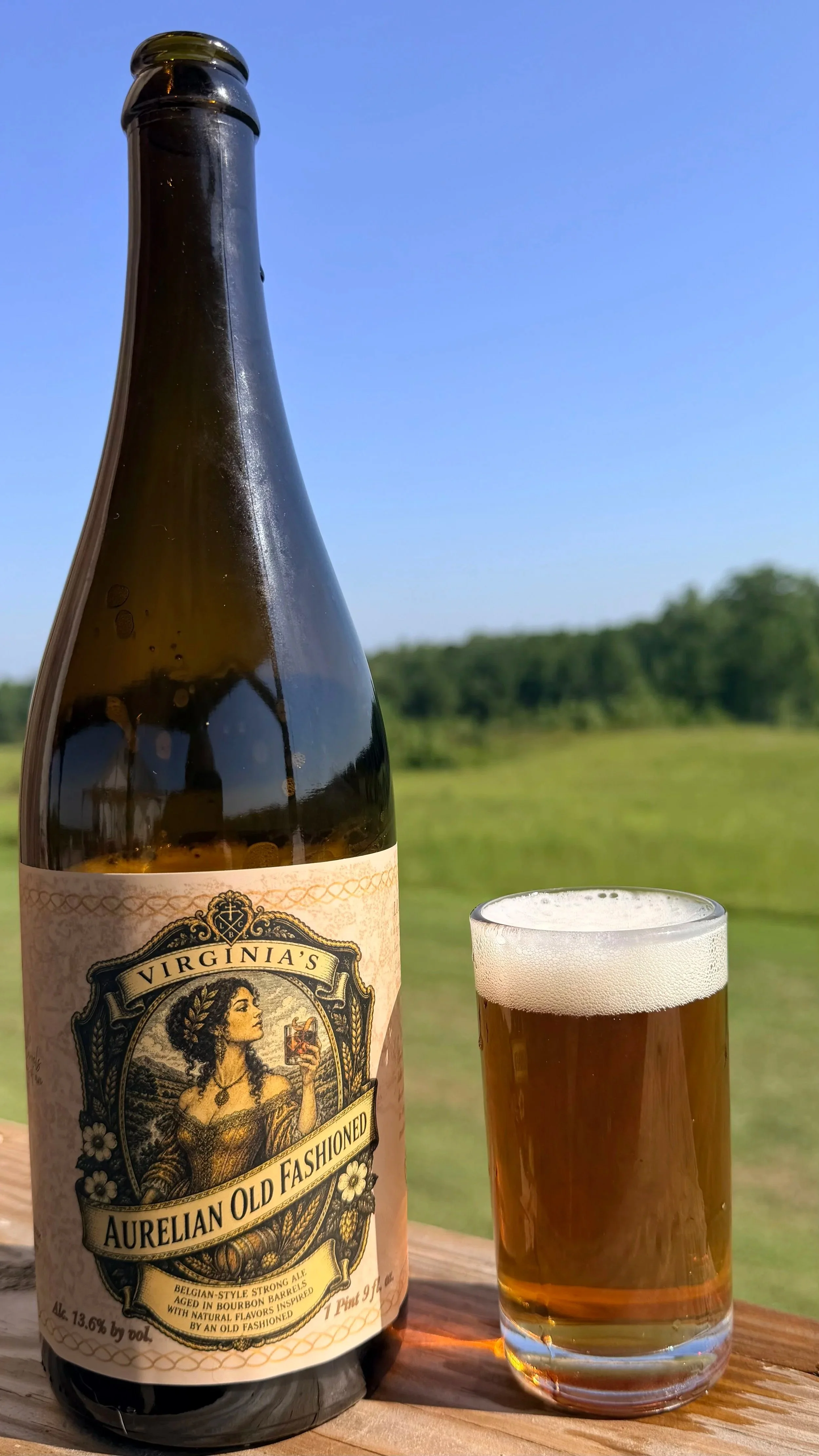 A bottle of beer labeled "Aurelian Old Fashioned" and a glass filled with beer on a wooden surface outdoors, with a background of green trees and a clear blue sky.
