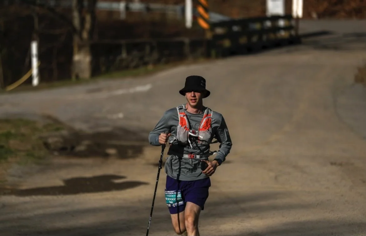 Trail runner wearing a hat and hydration vest, using trekking poles on a rural road.
