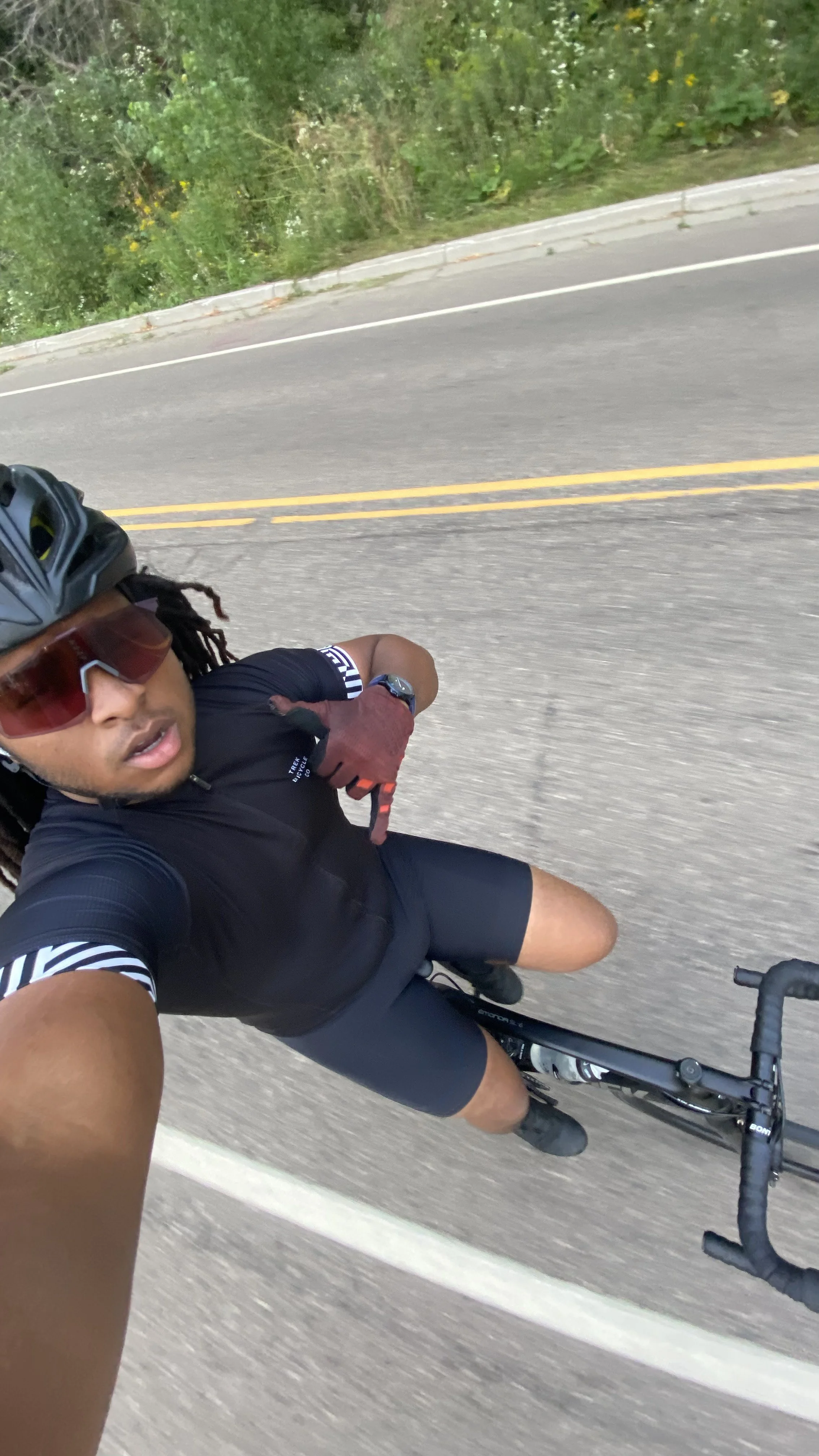 Cyclist taking a selfie while riding on a road with greenery in the background, wearing a helmet, sunglasses, and cycling gear.