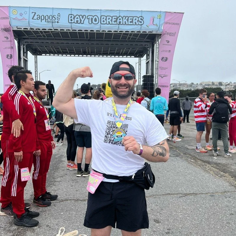 A participant poses with a medal at the "Bay to Breakers" event, featuring a banner overhead and a crowd in athletic attire.