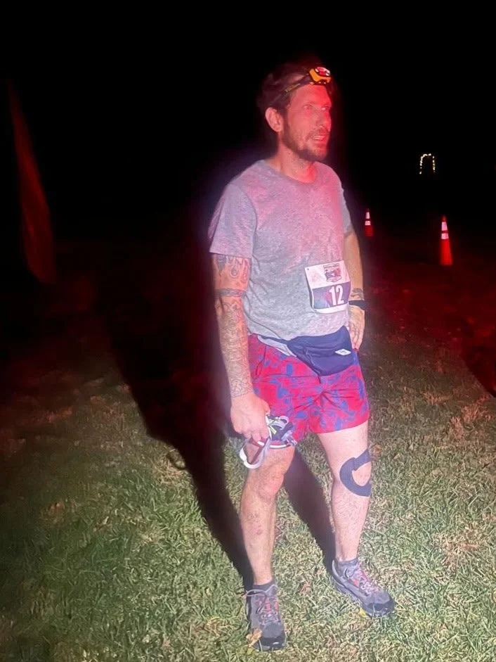 Man with backpack standing on grassy hill overlooking a large lake and distant mountains under a cloudy sky.