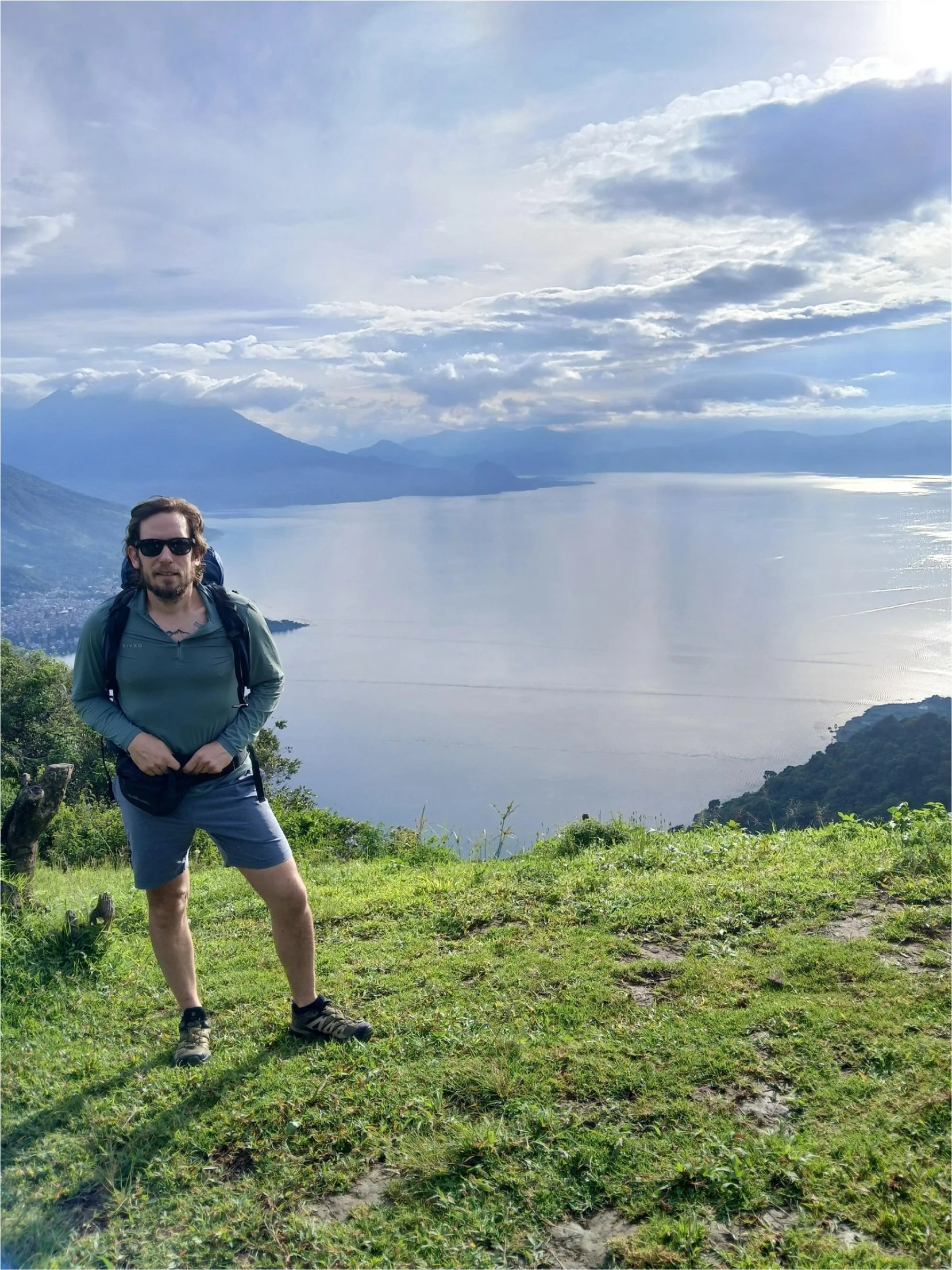 Man with backpack standing on grassy hill overlooking a large lake and distant mountains under a cloudy sky.
