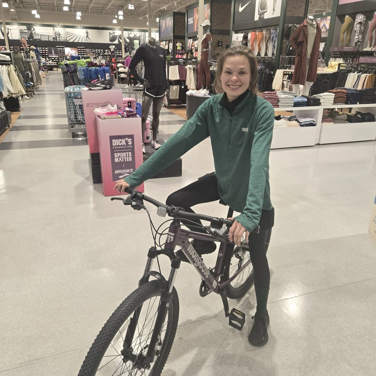 Person in green jacket posing with a mountain bike inside a sports store.