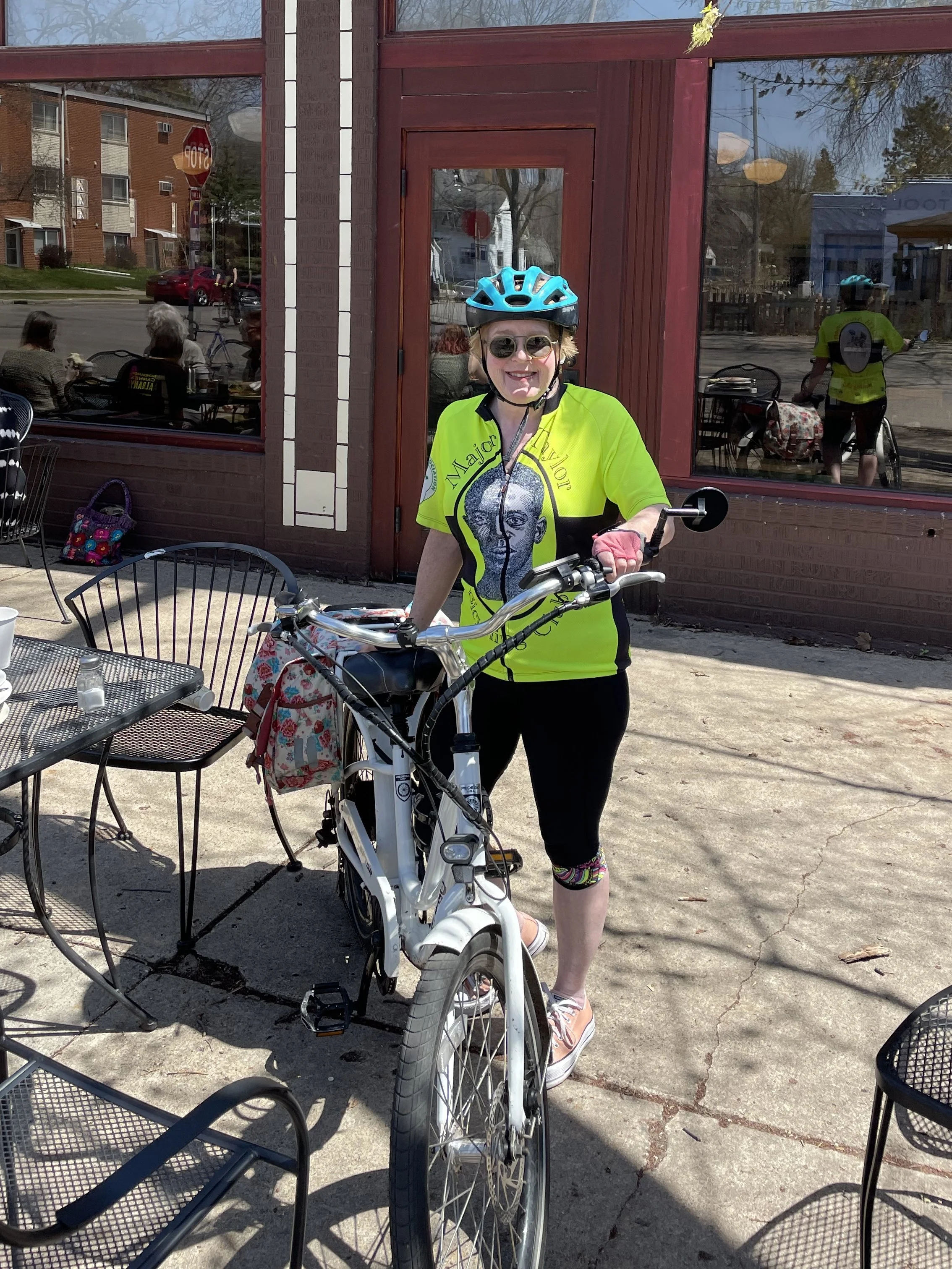 A person wearing a bright yellow cycling jersey and a blue helmet is standing outside a cafe with a white bicycle. They are smiling, and there are outdoor tables and chairs around them. The cafe has large glass windows.