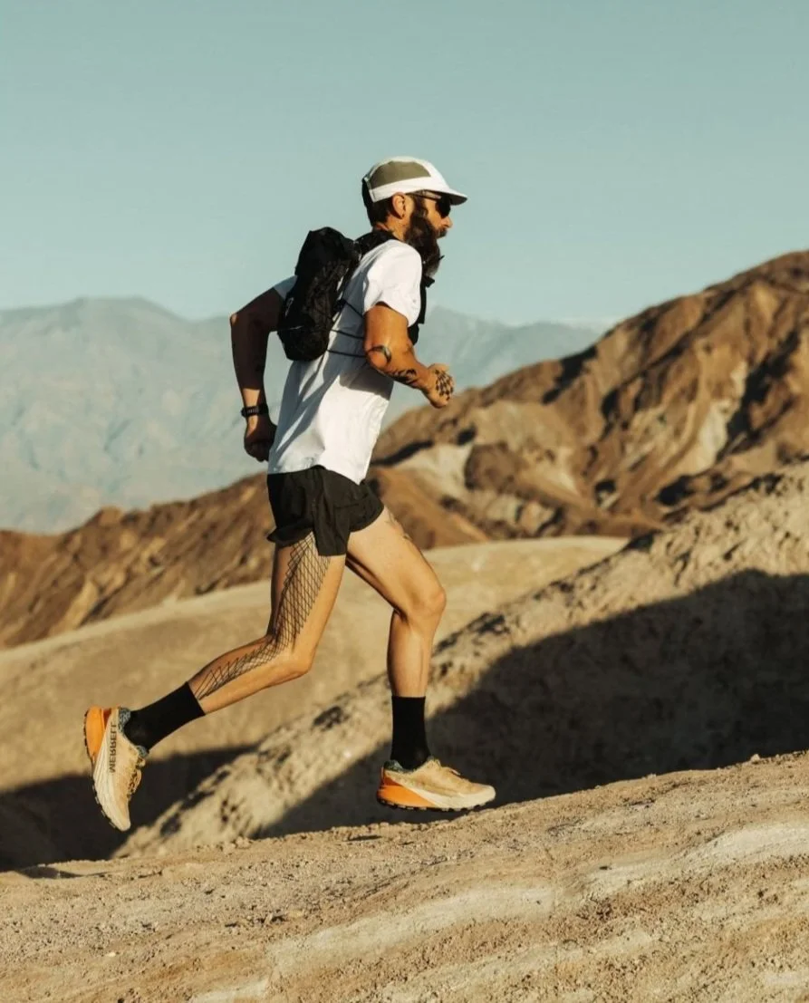 Man trail running in desert landscape, wearing a white shirt, black shorts, running shoes, sunglasses, cap, and backpack, with mountains in the background.