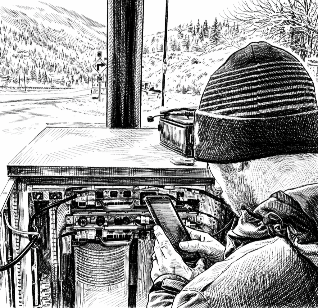 A person wearing a beanie and jacket is looking at their phone while working on outdoor electrical equipment near a roadside in a mountainous area with snow.