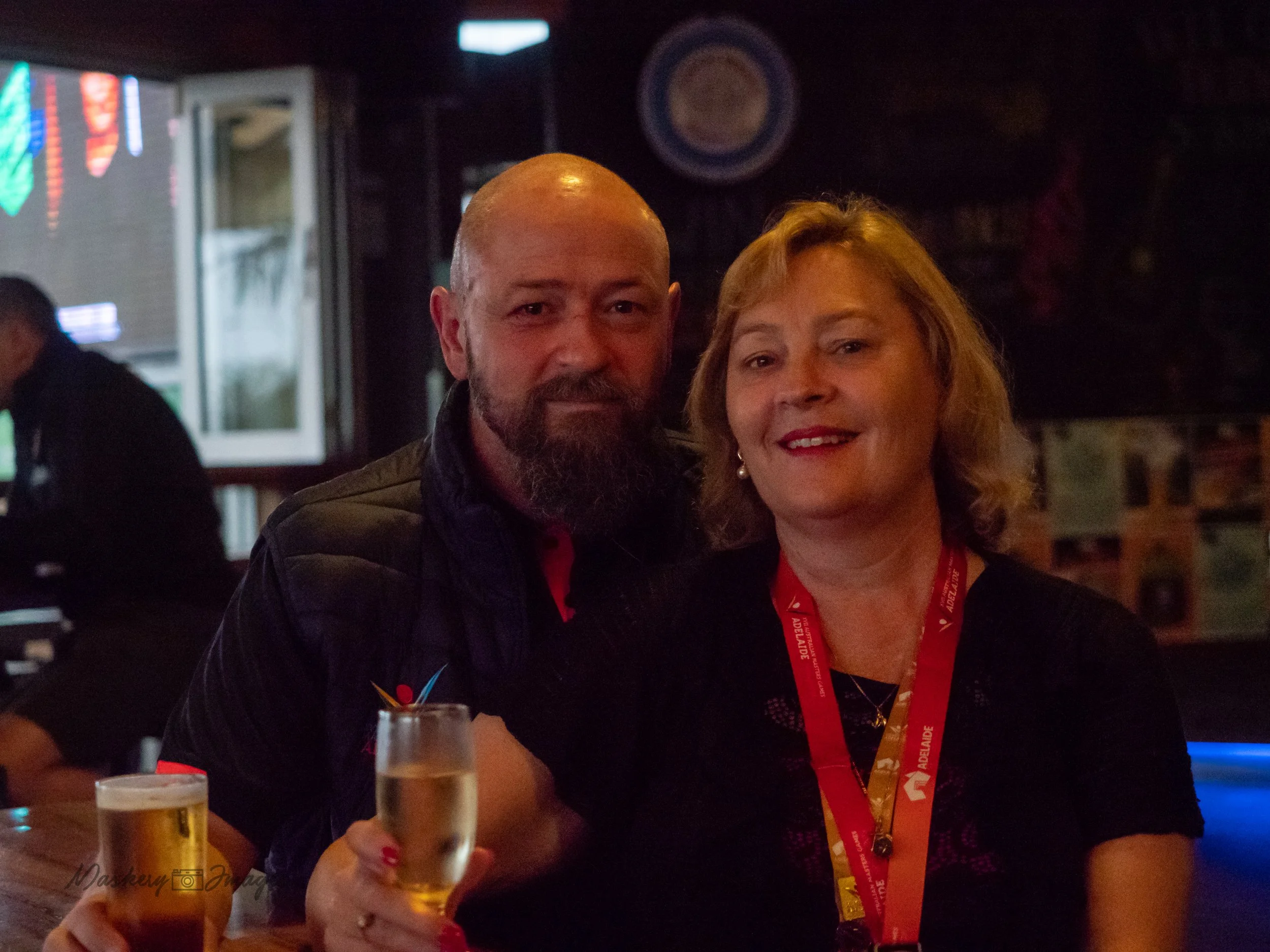 A man and woman posing together in a bar or pub, holding drinks and smiling at the camera. The woman is wearing a black top and a red lanyard, and the man is wearing a dark jacket.